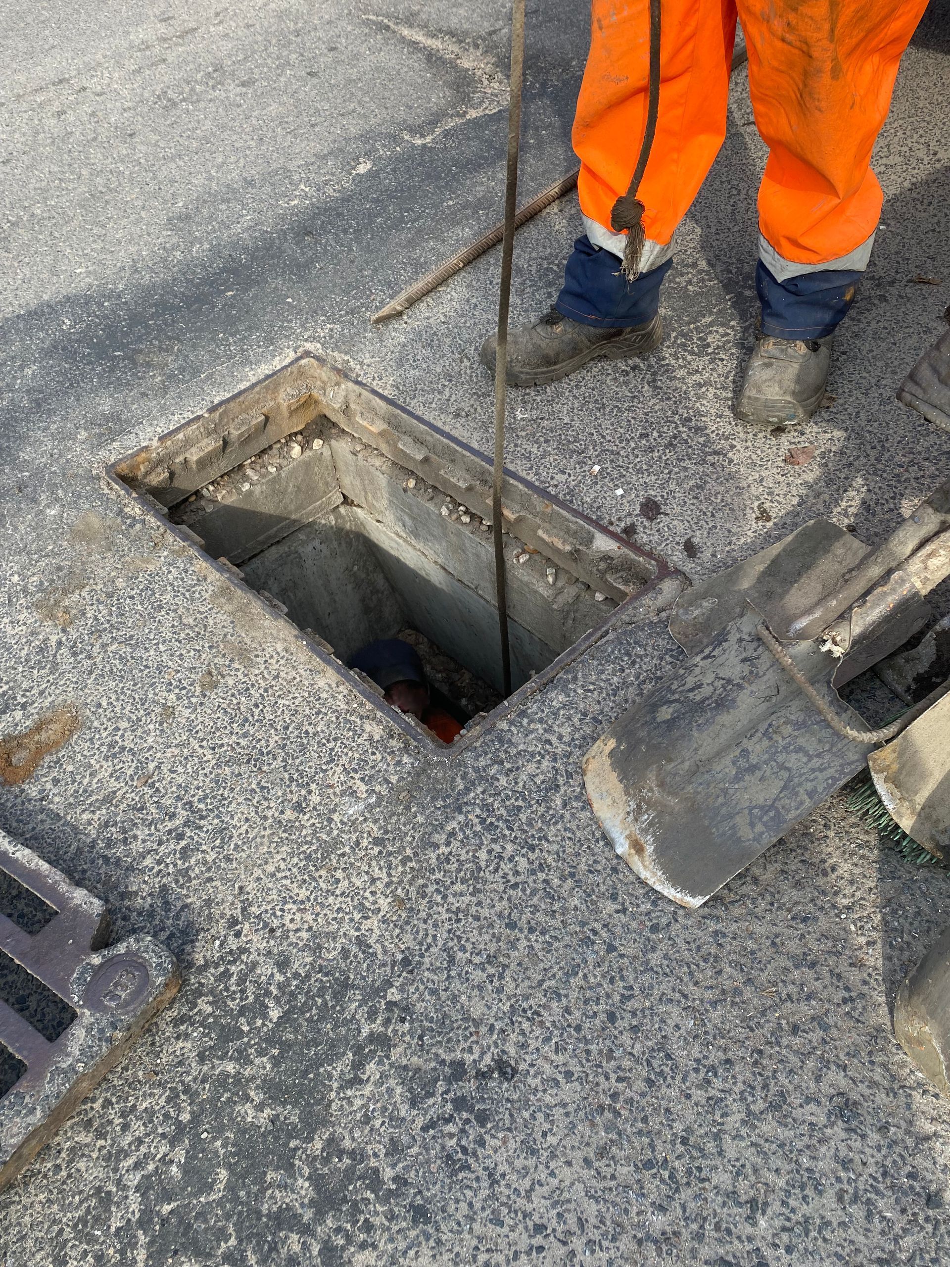 Person in orange workwear near open manhole; grey road surface.