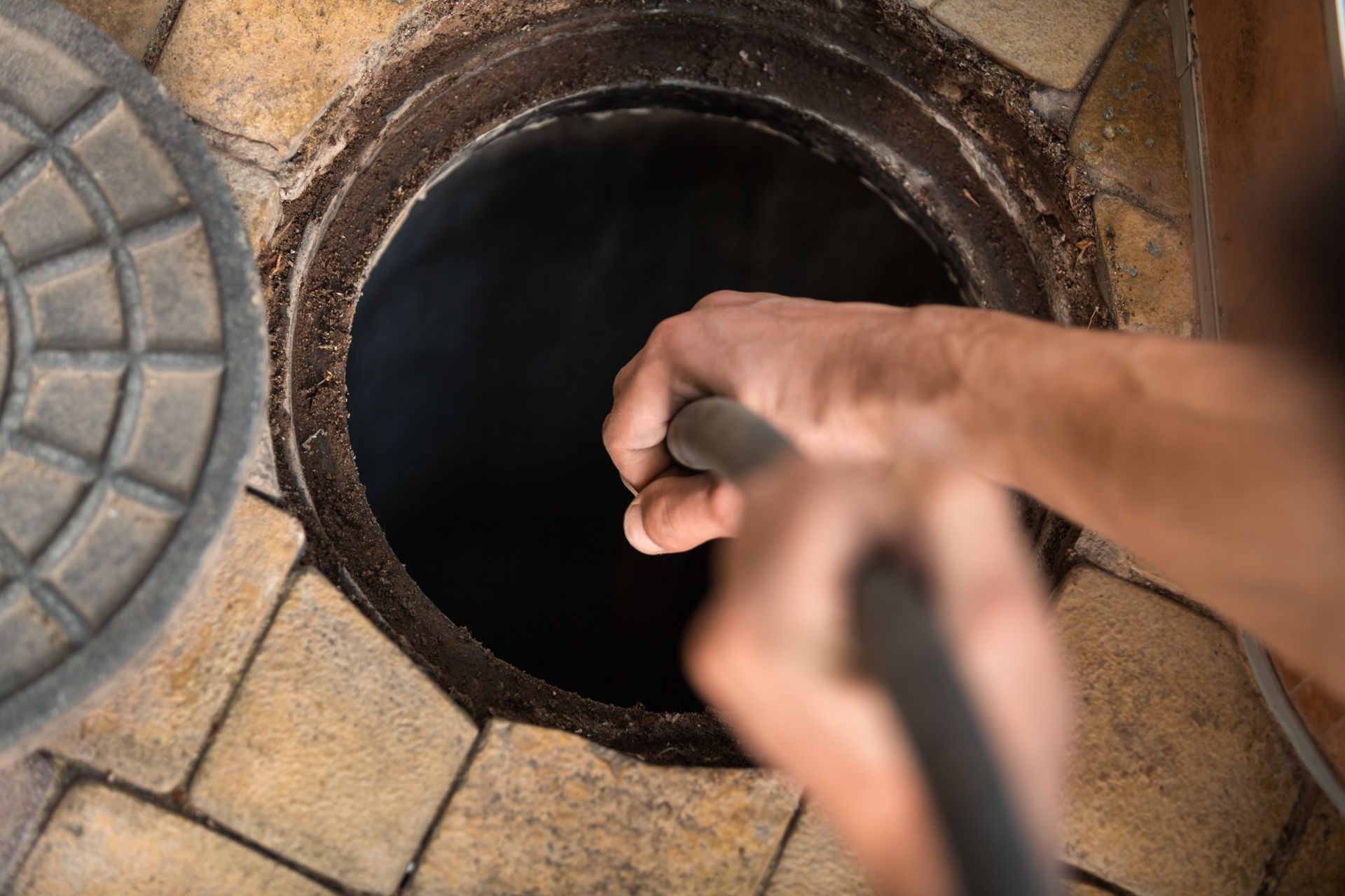 Person's hands holding a hose inside an open brick-lined access point, possibly a well or inspection chamber.