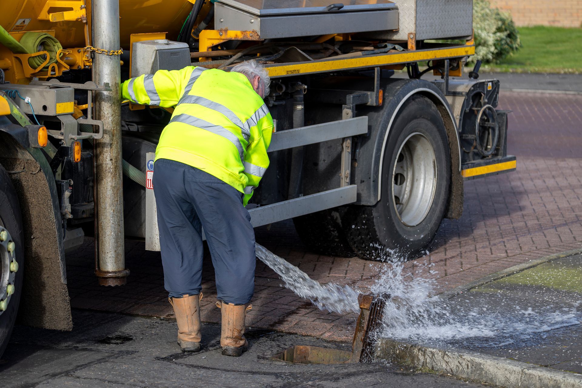Worker in high-vis vest flushing a street drain with water from a large yellow truck.