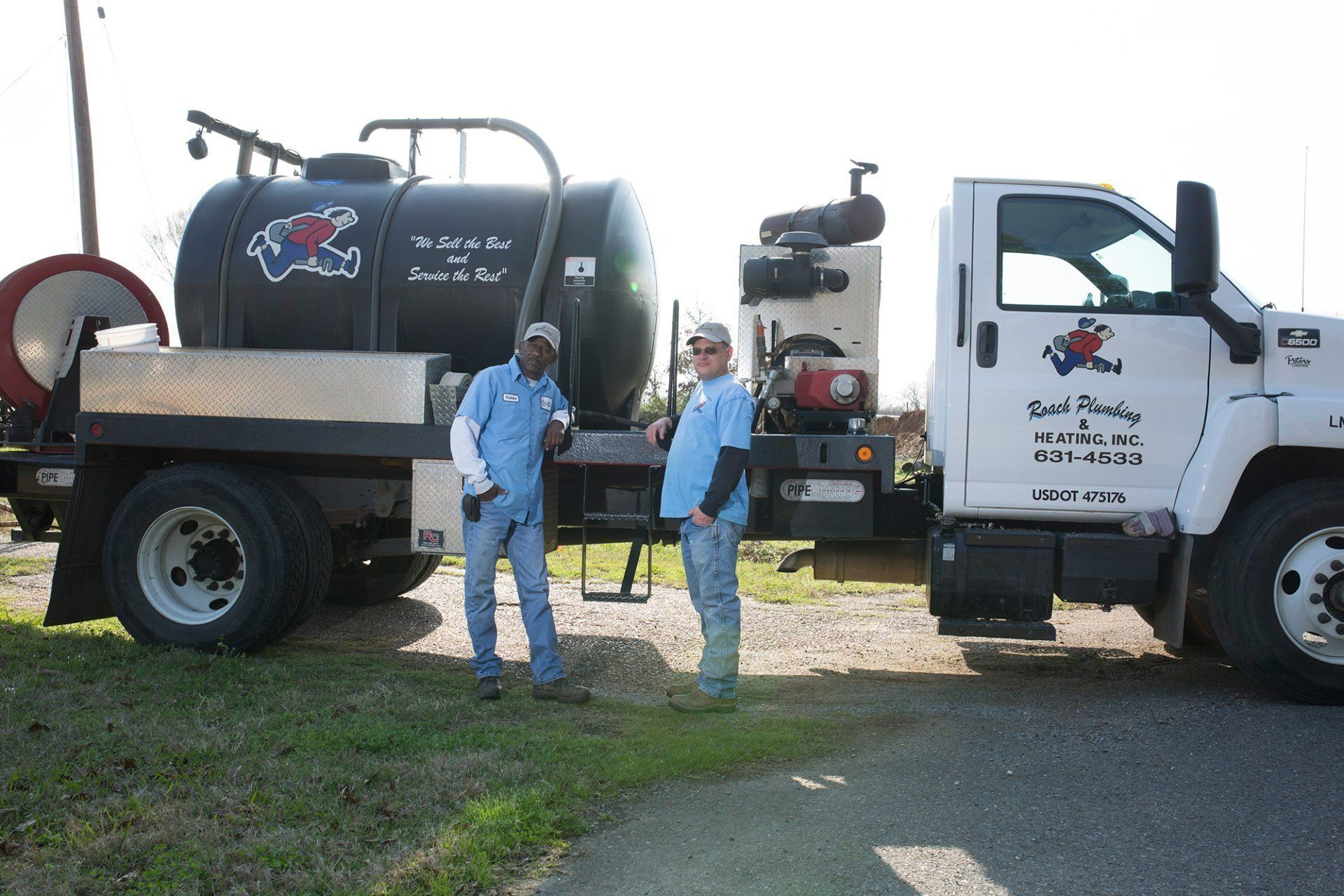 Two people stand near a black tanker truck with a business logo in a field.