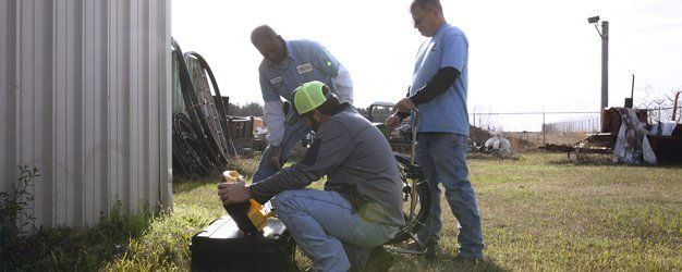 Three people working in a grassy outdoor area near a corrugated metal building.