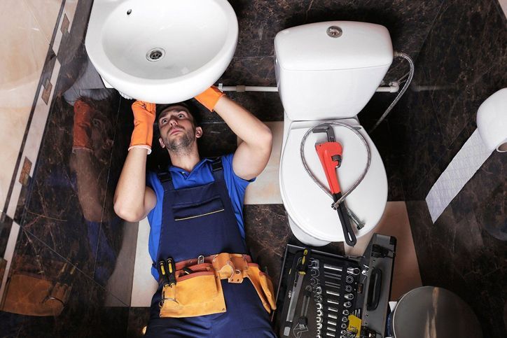 Plumber in blue coveralls fixing a sink in a bathroom, with a toolbox nearby. Orange gloves on.