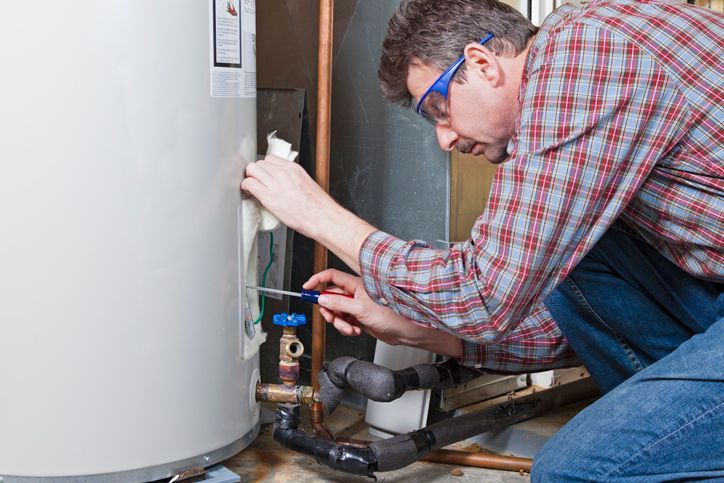 Man in plaid shirt and safety glasses working on a water heater, crouched in a utility room.