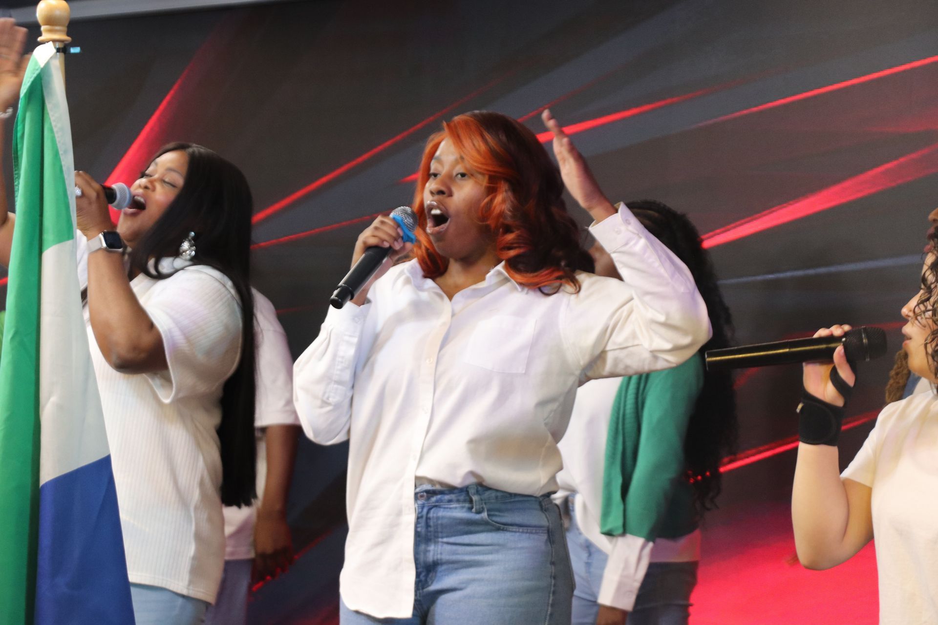 Singing group with a flag. Woman with orange hair sings into microphone, wearing a white shirt and jeans.
