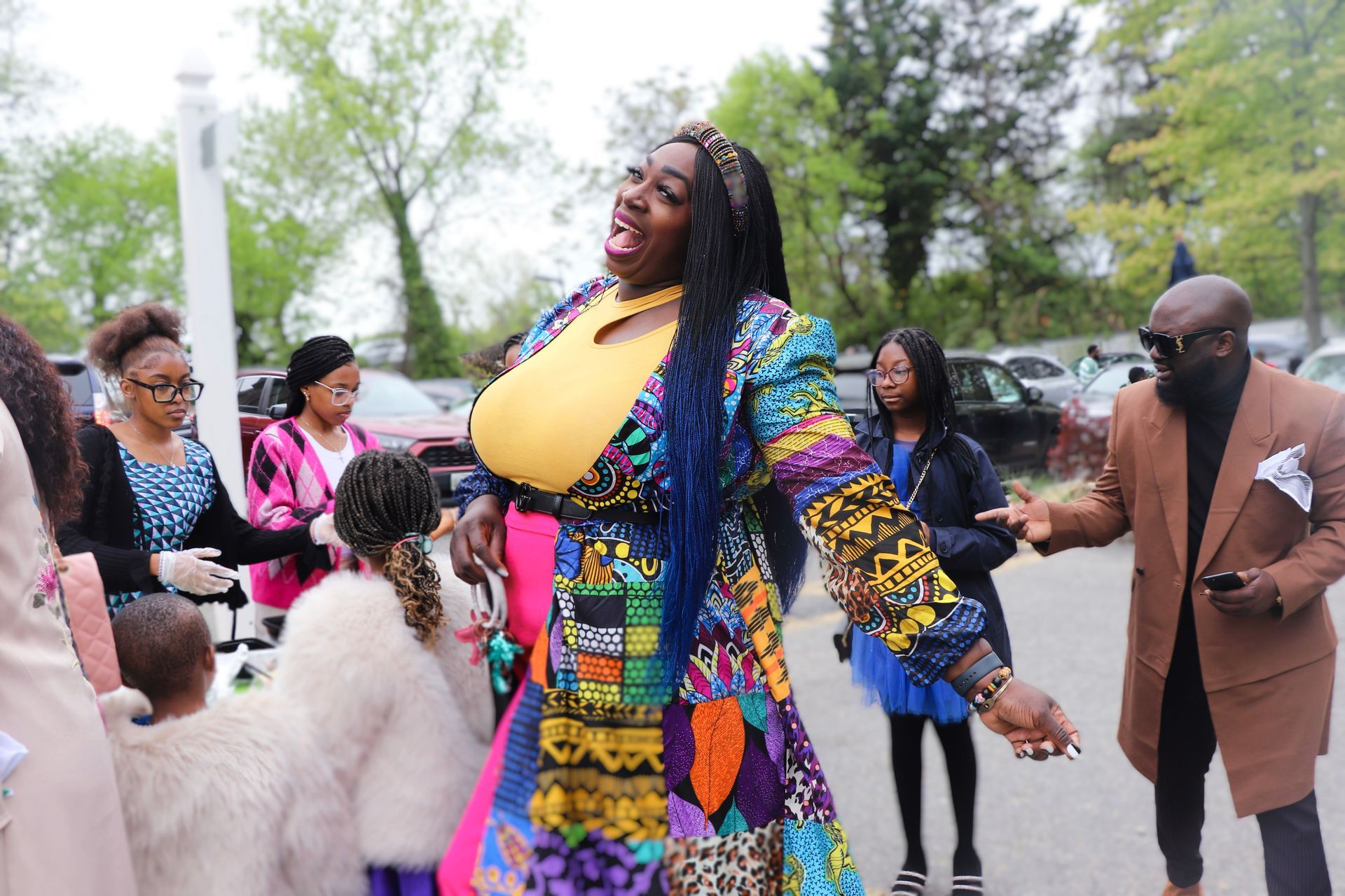 Woman in vibrant patchwork coat and yellow top smiles, surrounded by people outdoors.