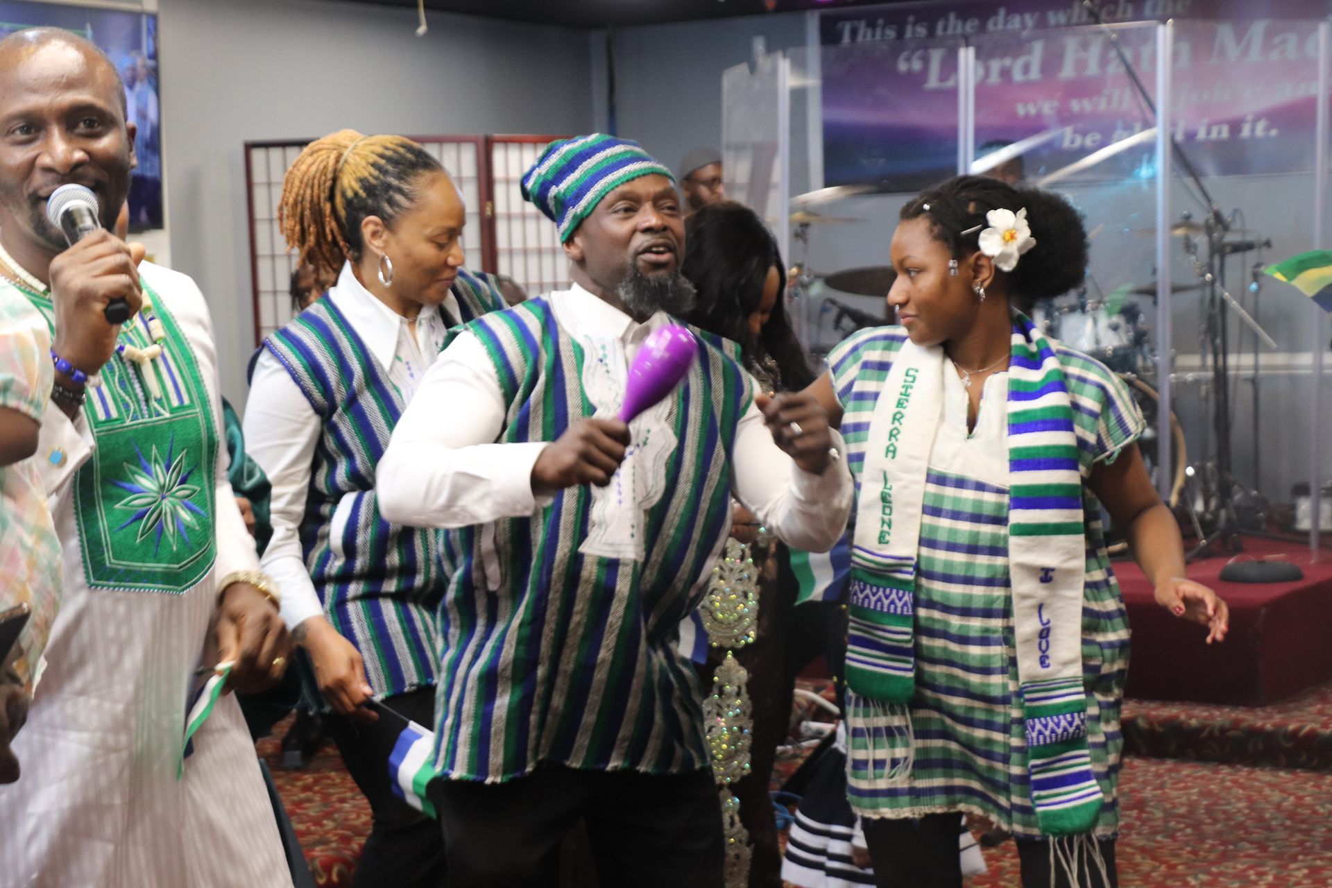 People in matching green and white outfits, dancing in what appears to be a church, holding instruments and smiling.