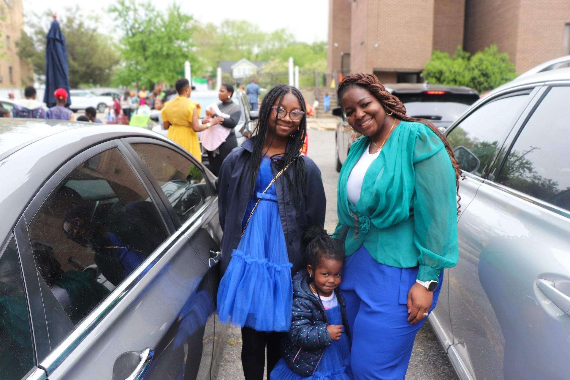A family of three smiling, standing between parked cars outdoors; all wearing shades of blue.