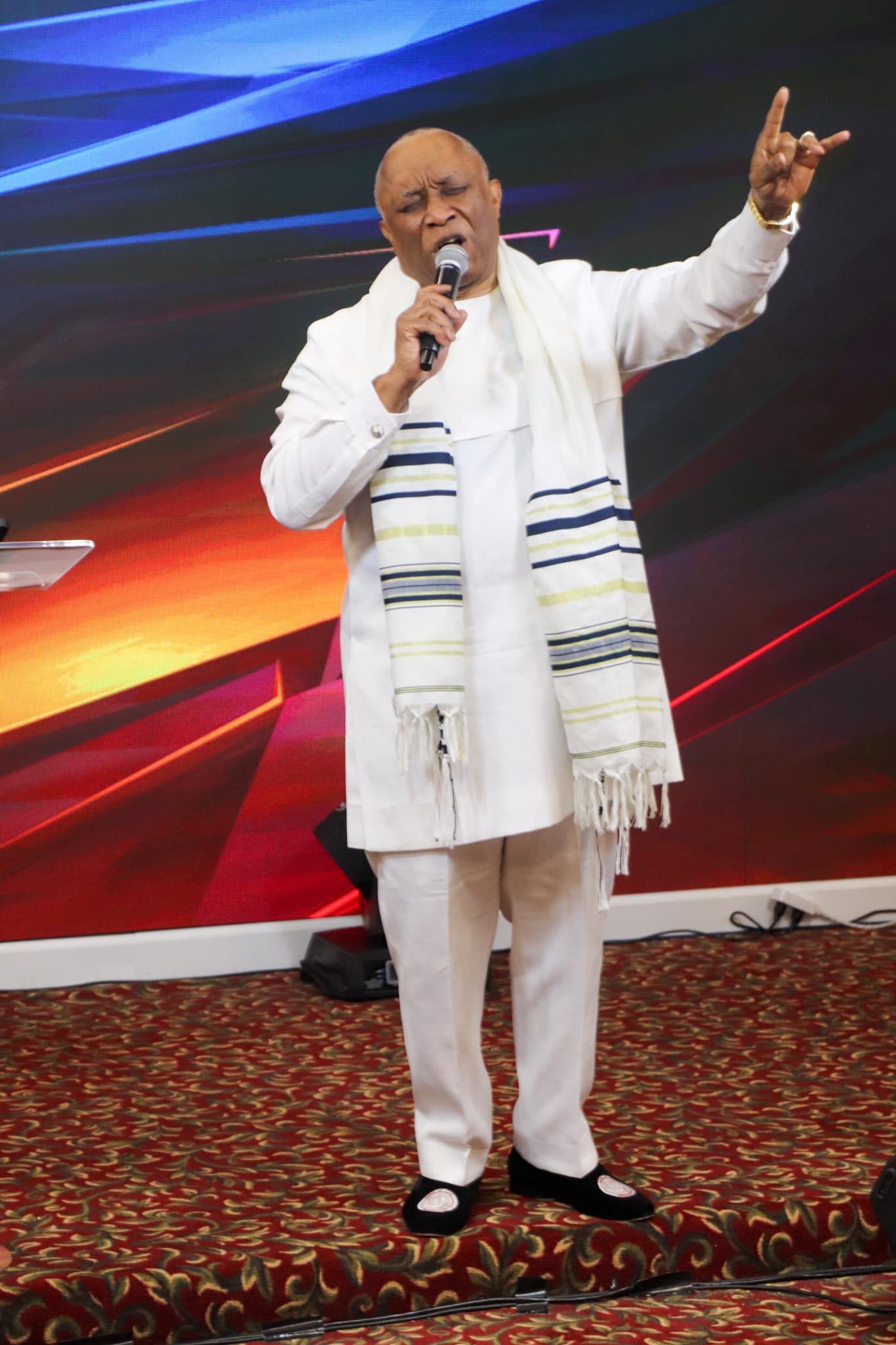 Man in white suit with scarf, raising hand. Standing on a stage with colorful backdrop.