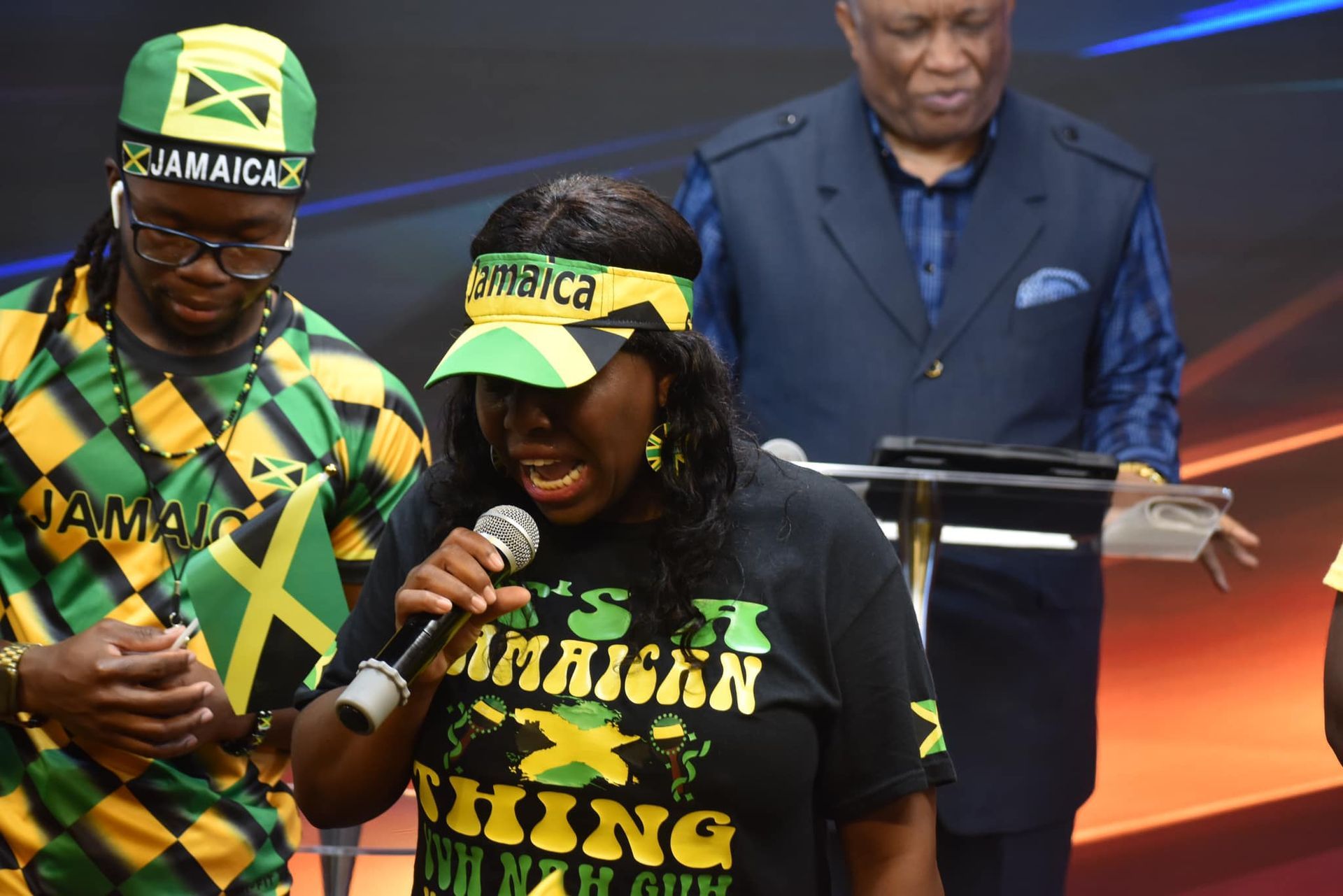 A woman singing into a microphone with Jamaican flag clothing. Man with a hat and flag, and a man behind a podium.