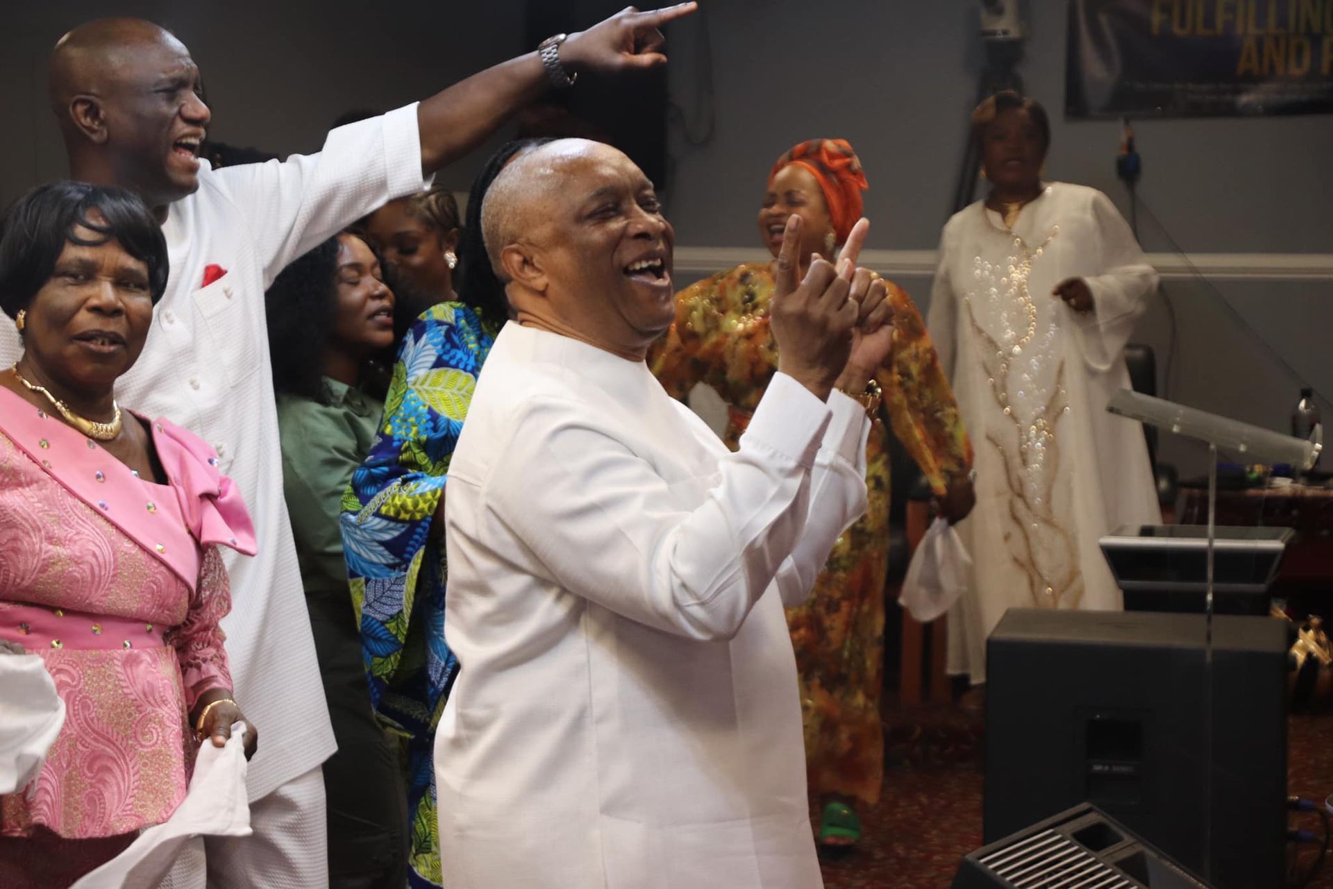 Group of people in a room, some singing and gesturing. Man in white suit is center. Others wear colorful outfits.
