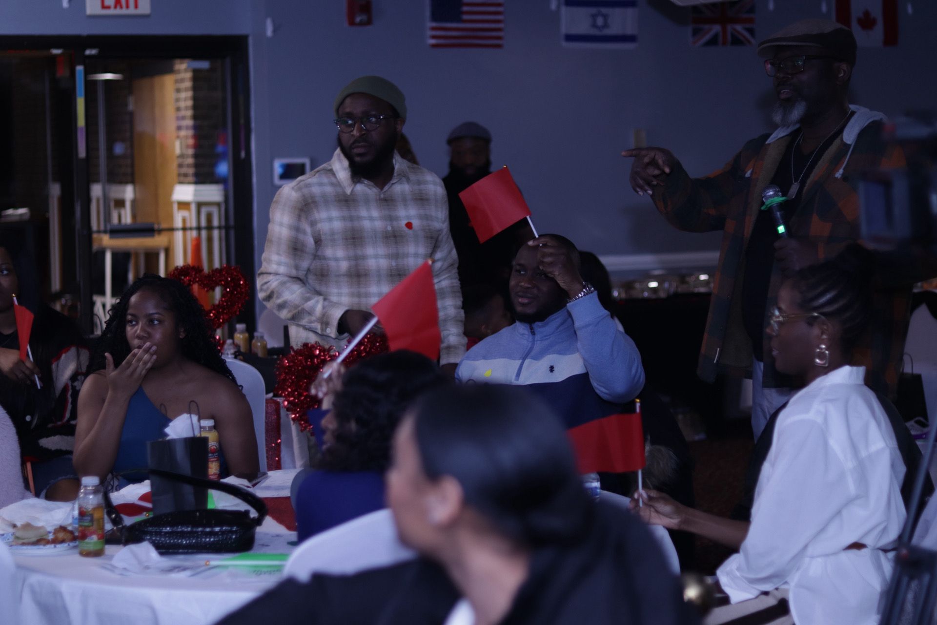People at a gathering, some holding red flags, a man gestures. Indoors, lights, tables, and flags.