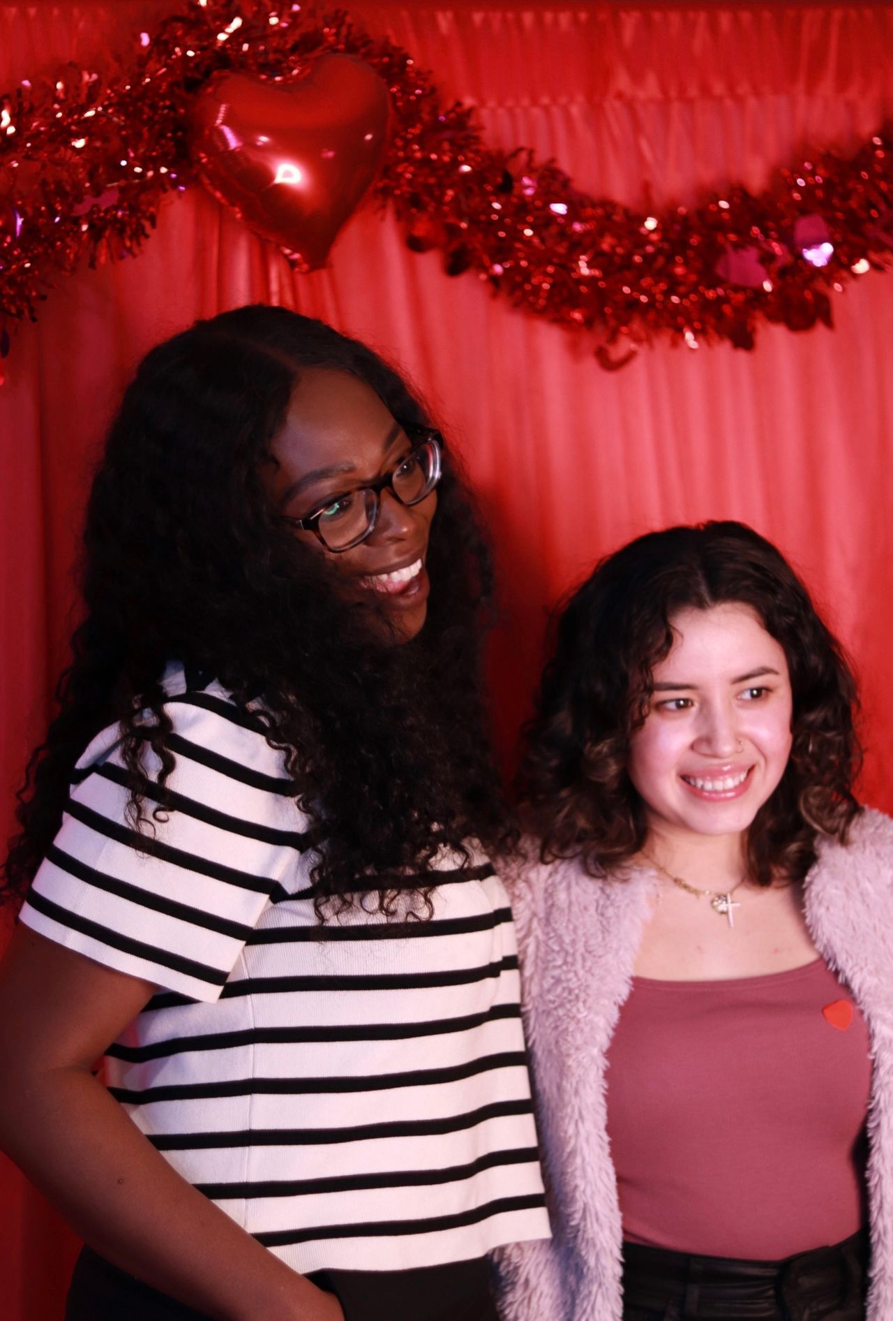 Two women smiling in front of a red Valentine's Day backdrop; one wearing glasses and a striped shirt, the other, pink.