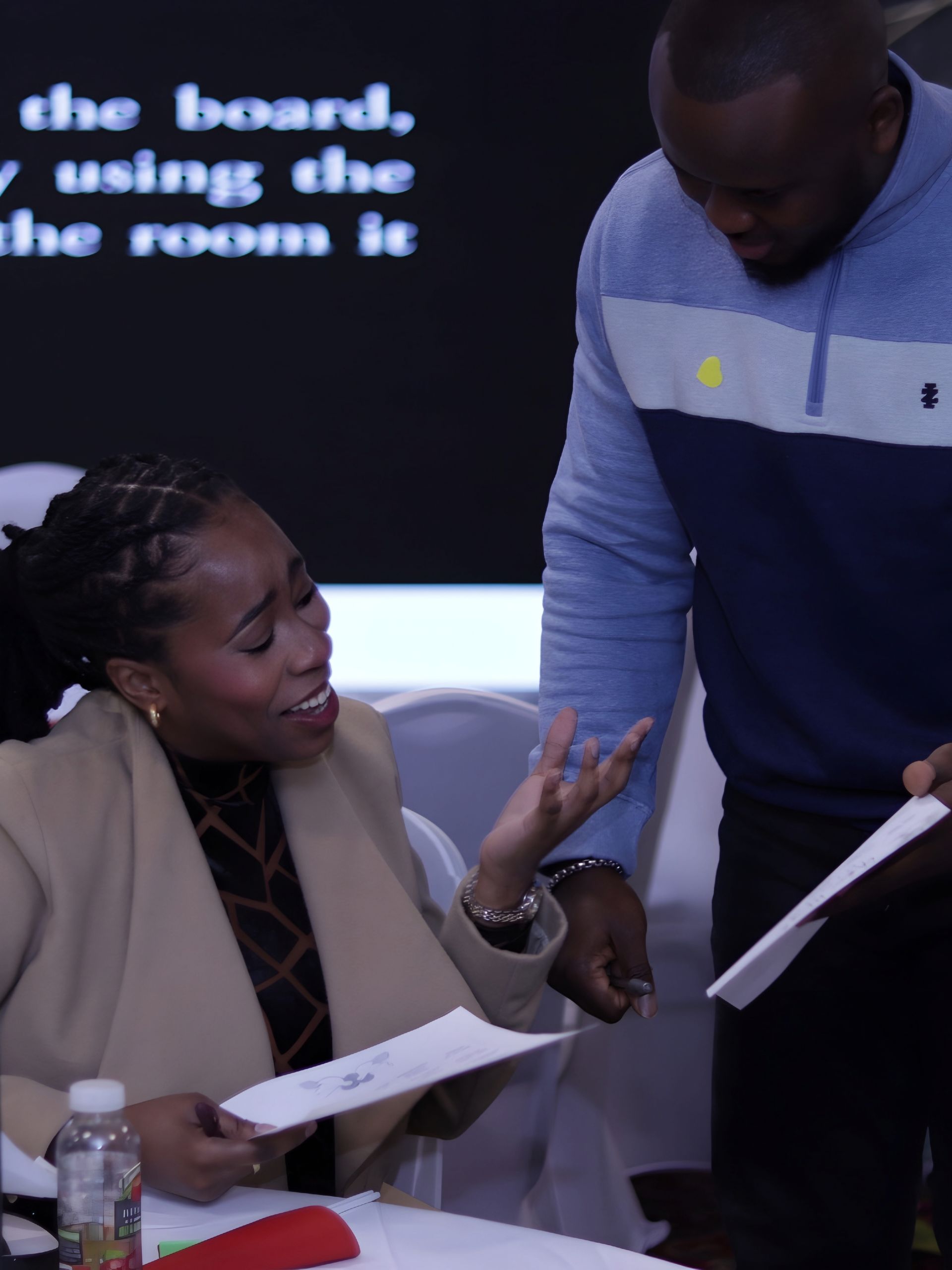Woman gestures, holding papers, talking to a man in a meeting room, both looking at documents.