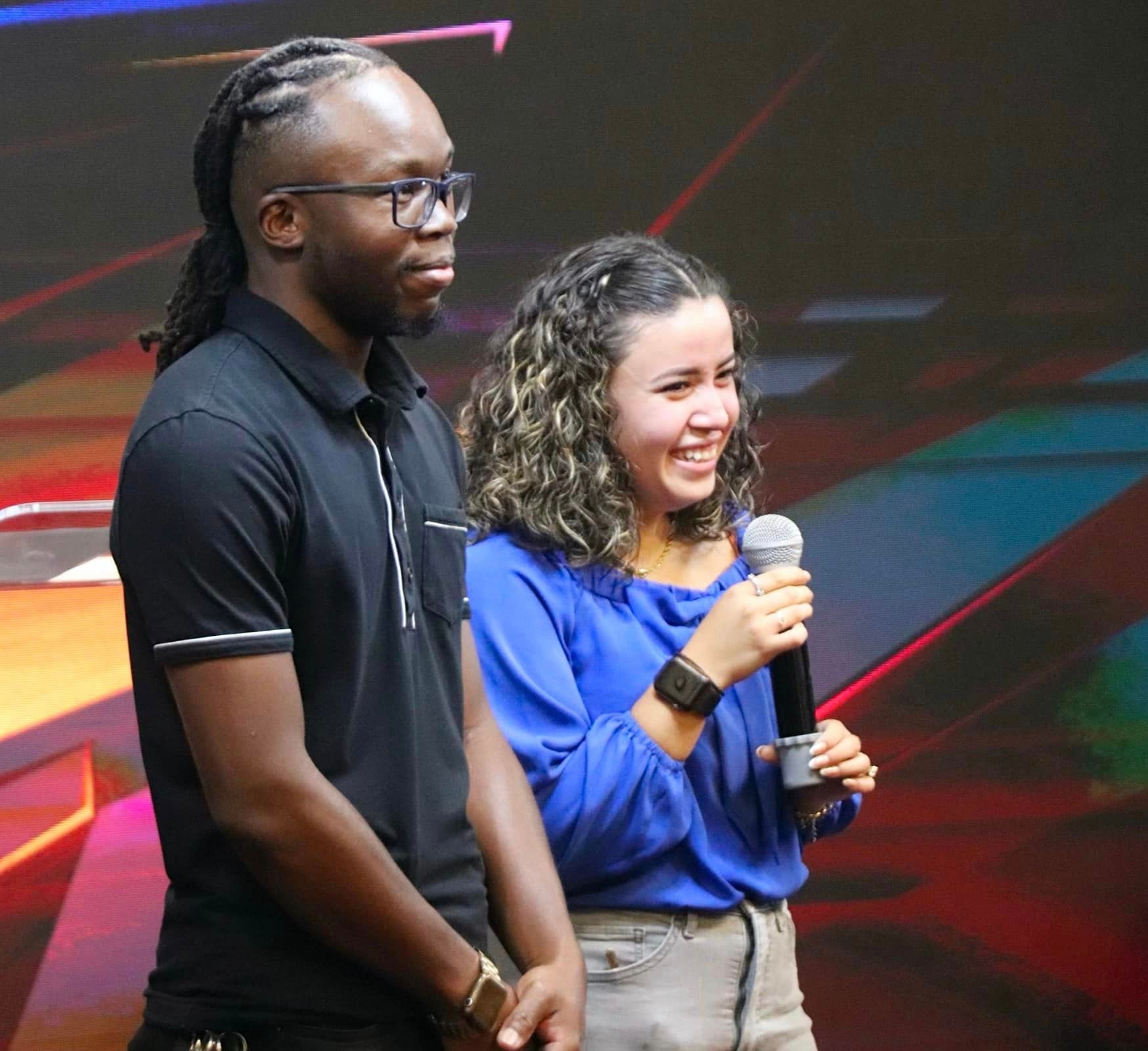 Man with dreadlocks and glasses next to a smiling woman holding a microphone; onstage.