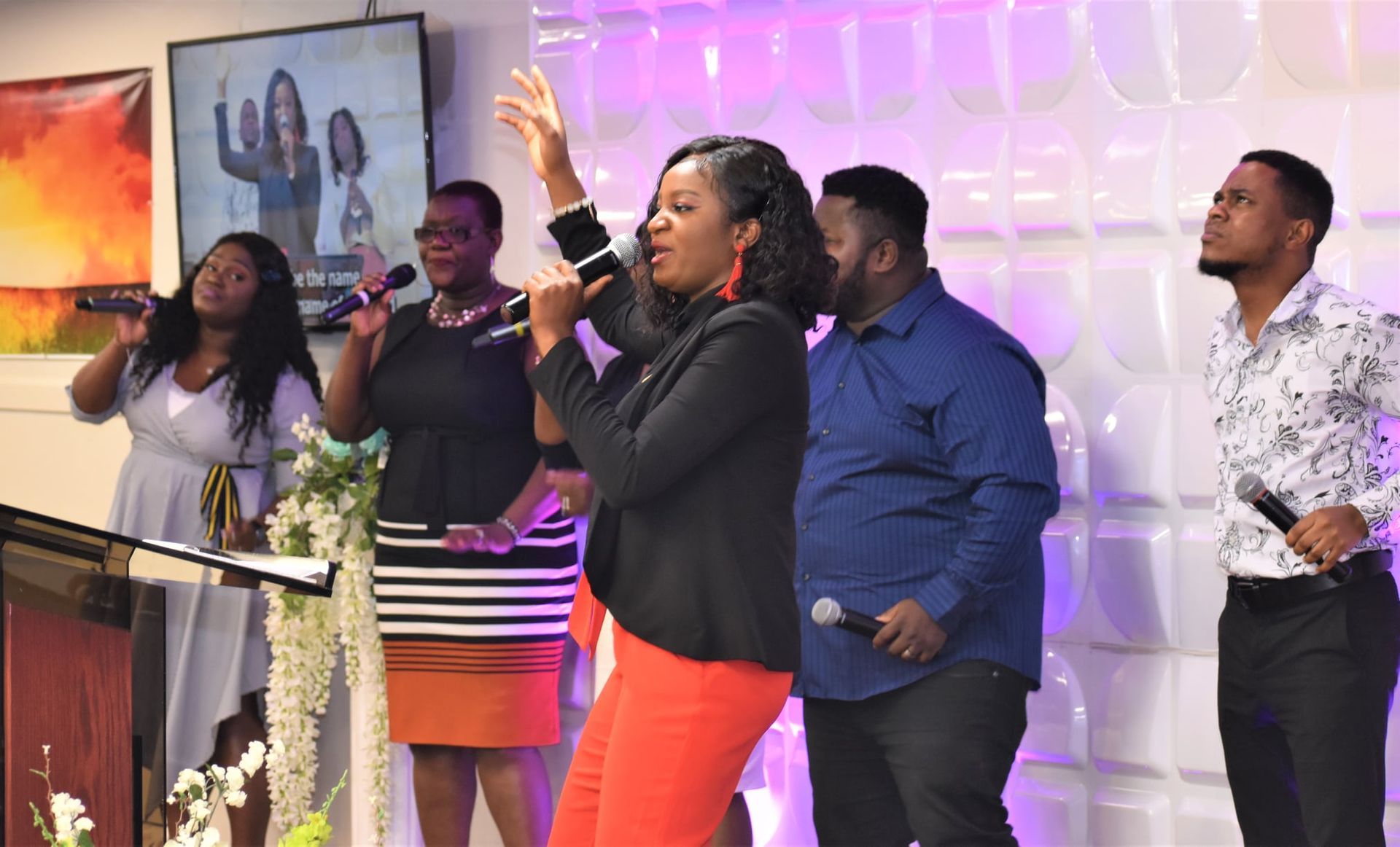 Choir singing on stage with microphones, raised hands.  Orange and white backdrop, diverse group.