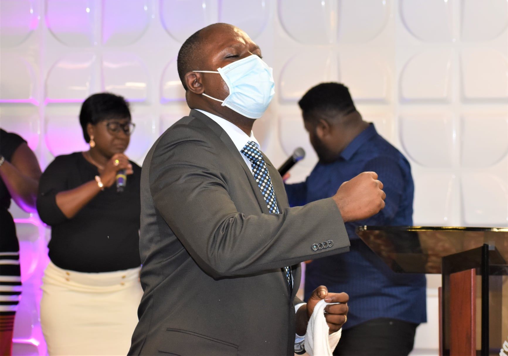 Man in suit with mask sings, eyes closed, in front of a white textured wall, with people in the background.