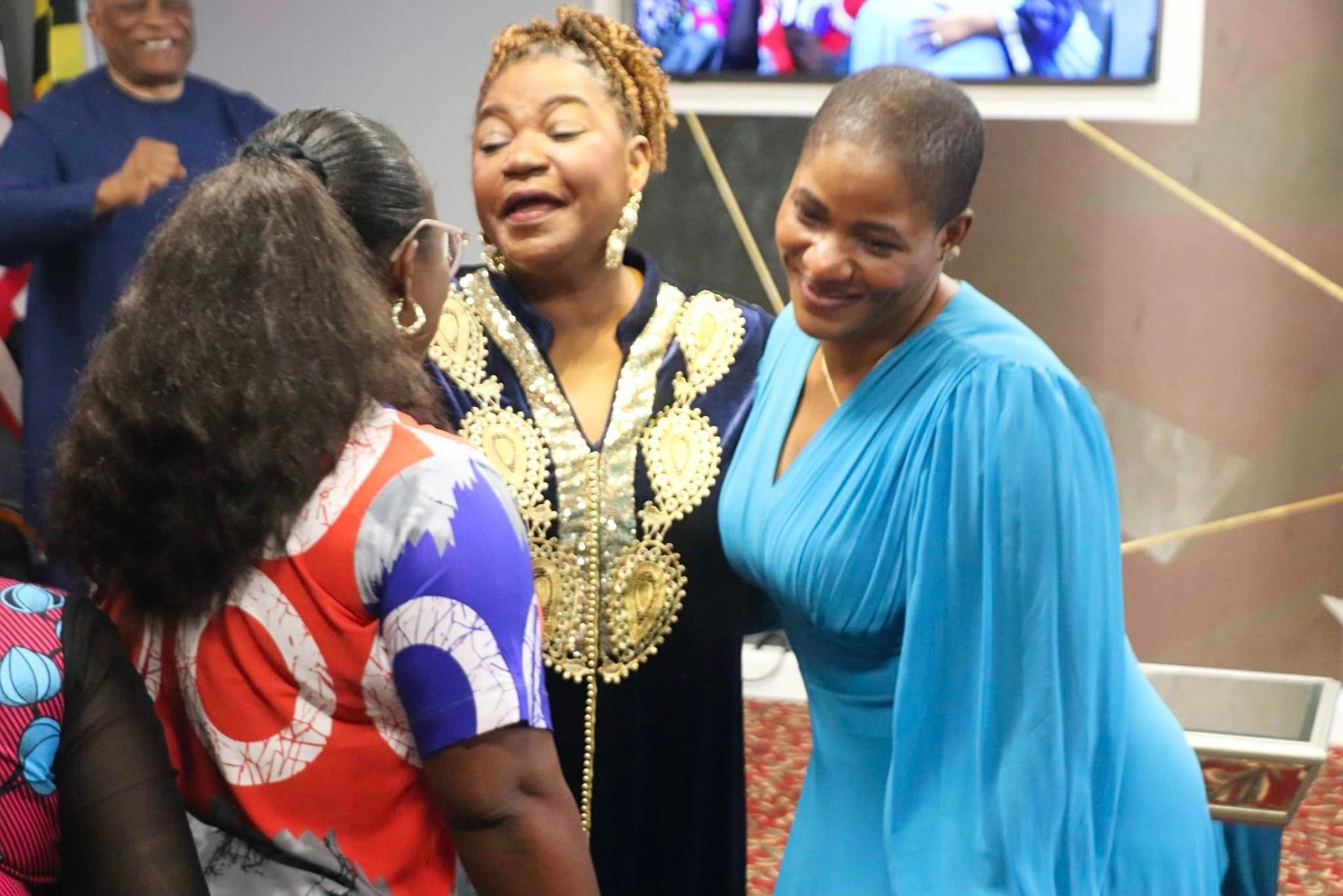 Three women in a room, embracing. One in blue dress, one in black and gold embroidered robe.