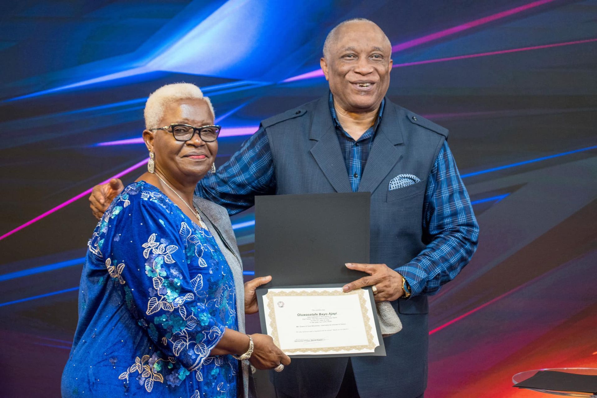 An older Black woman receives a certificate from an older Black man. They smile in front of a blue and purple background.