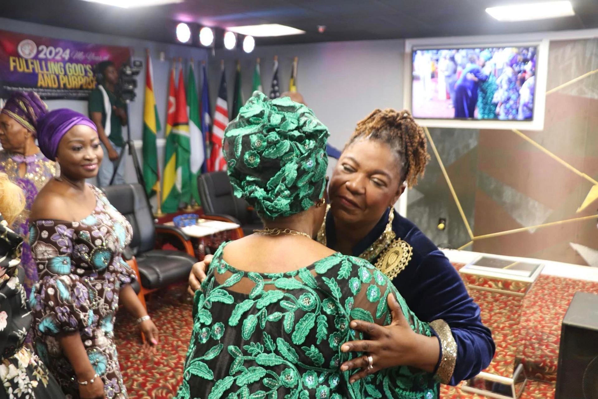 Women embrace in a room with flags and a monitor. One wears a green headwrap and patterned dress.