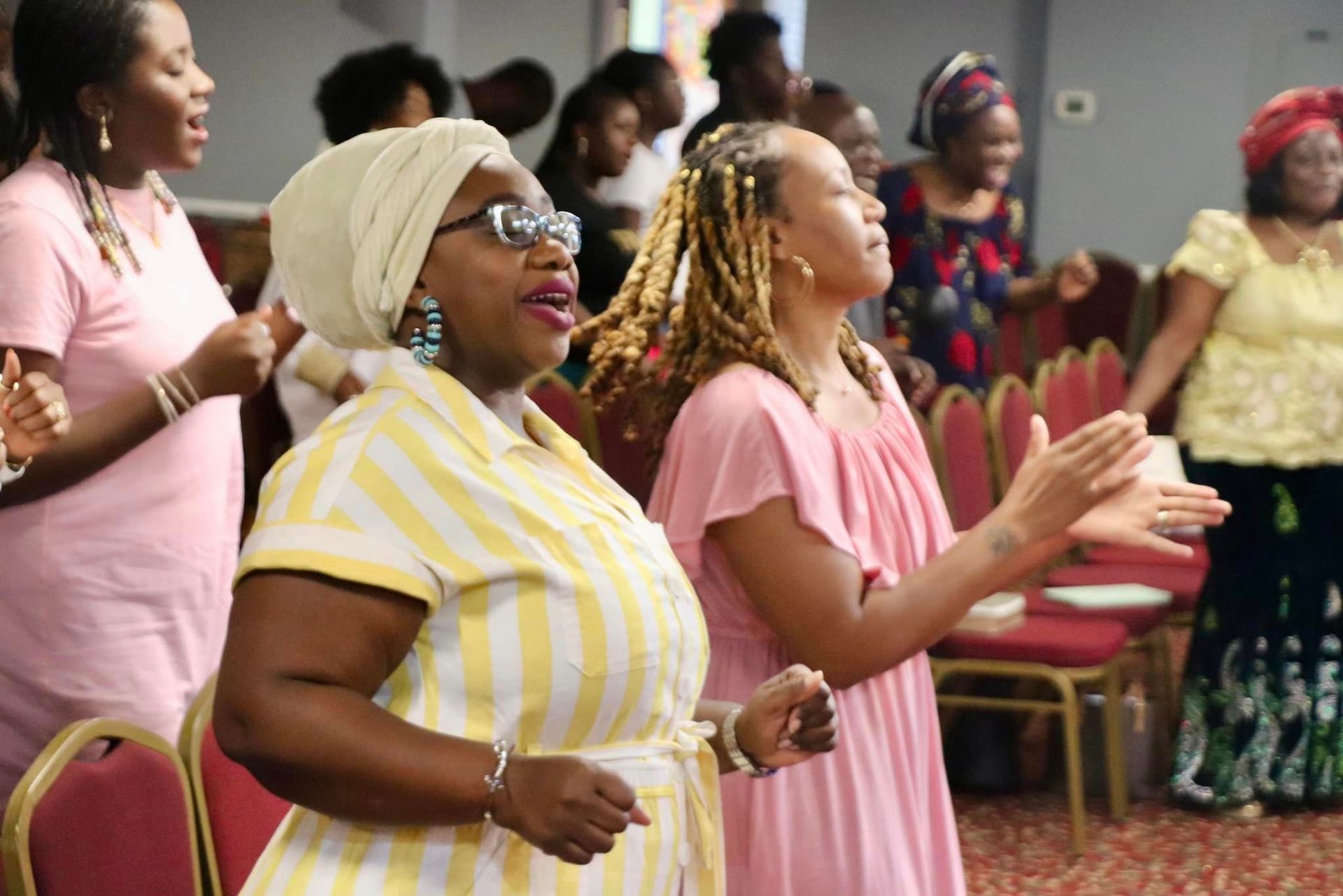 Group of women singing and clapping, expressions of joy, in a room with red chairs.