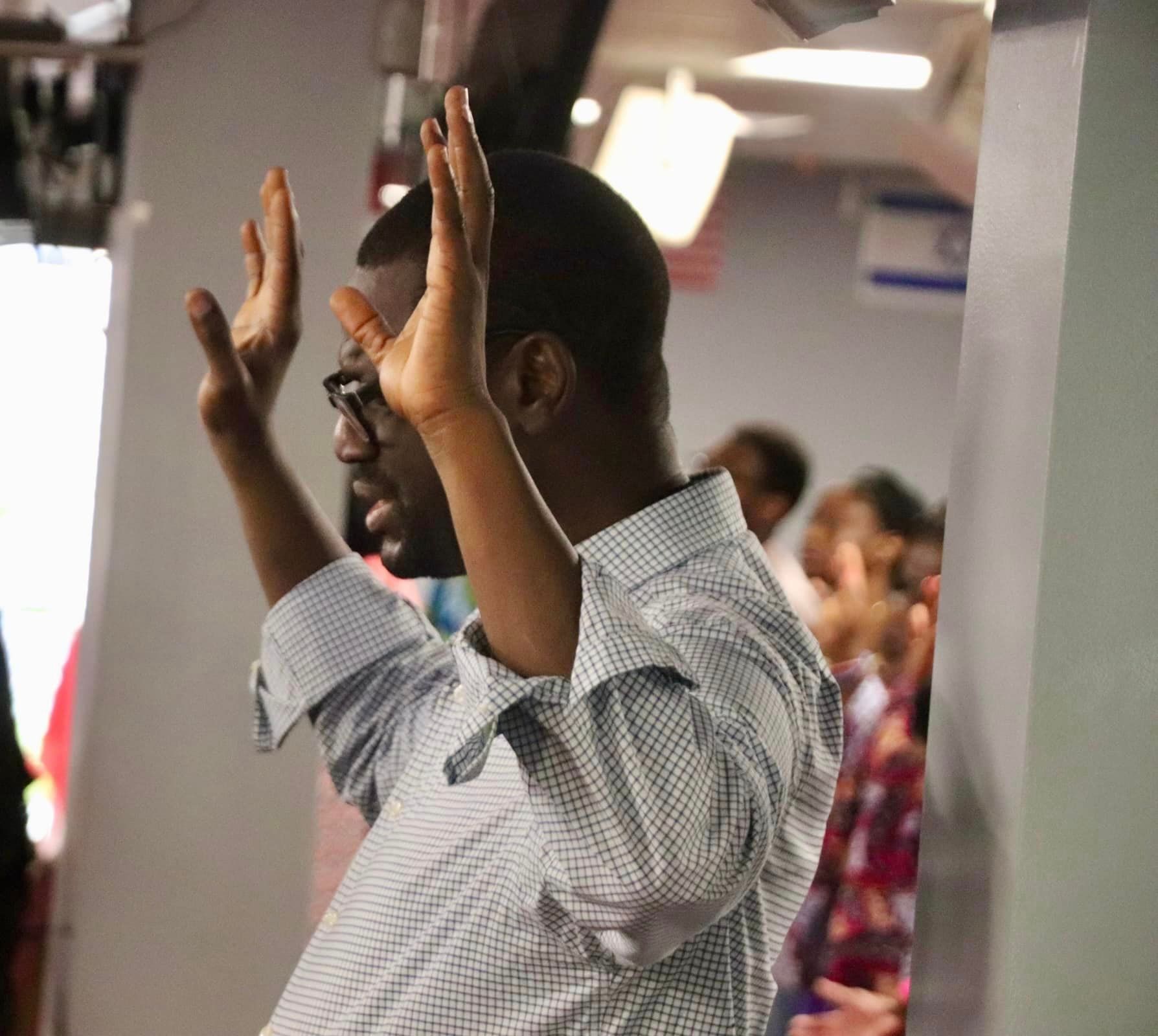 Man with hands raised in worship, wearing glasses and a light blue shirt, in a brightly lit church setting.