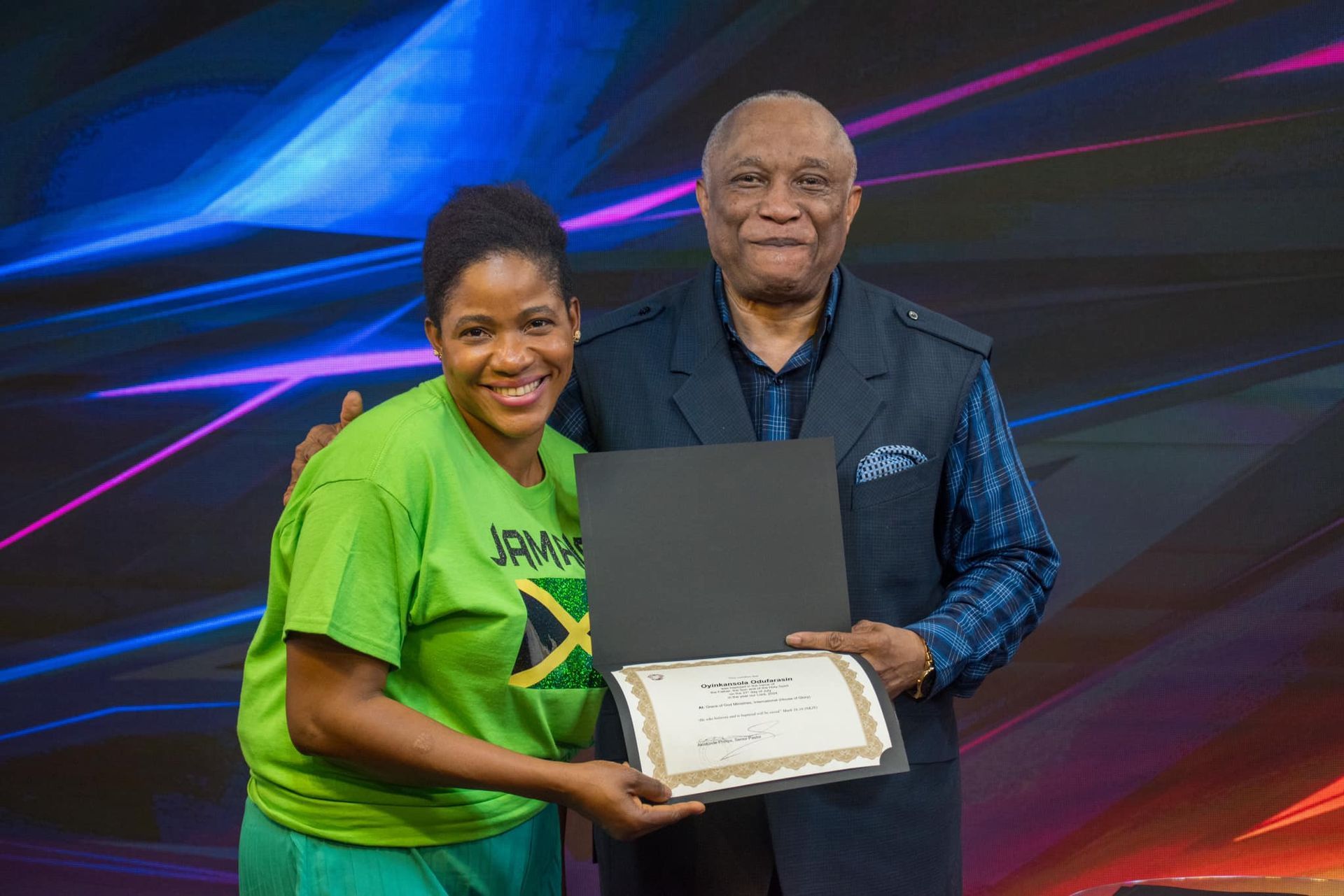 Woman in green shirt with Jamaican flag is presented with certificate by older man. They are smiling.