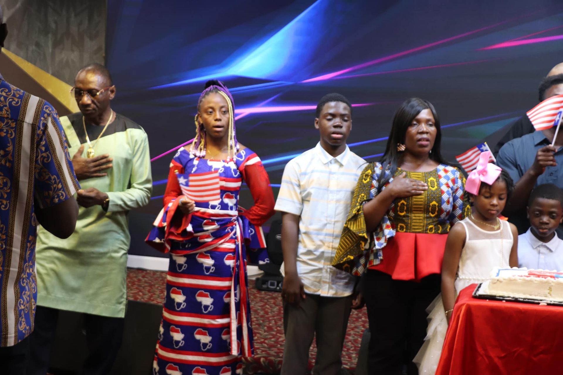 Group of people standing, holding American flags, hands on their hearts. Stage setting with a book and a screen.