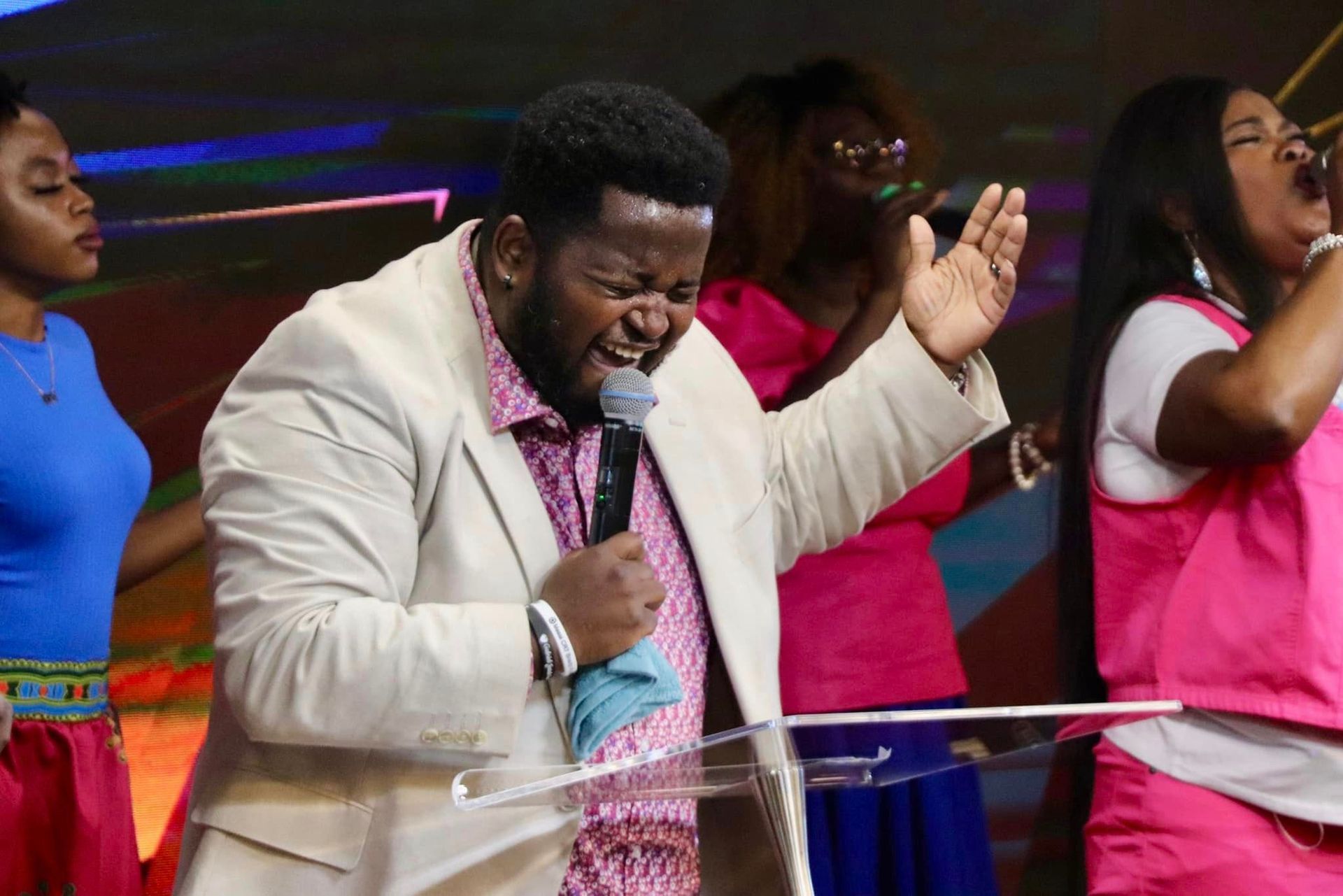 Man singing with raised hand at a podium, backed by a choir in a colorful setting.