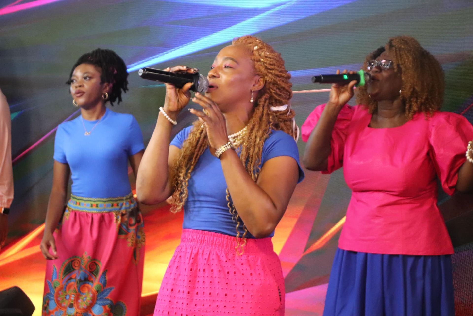 Three women singing into microphones on stage, wearing blue and pink outfits.