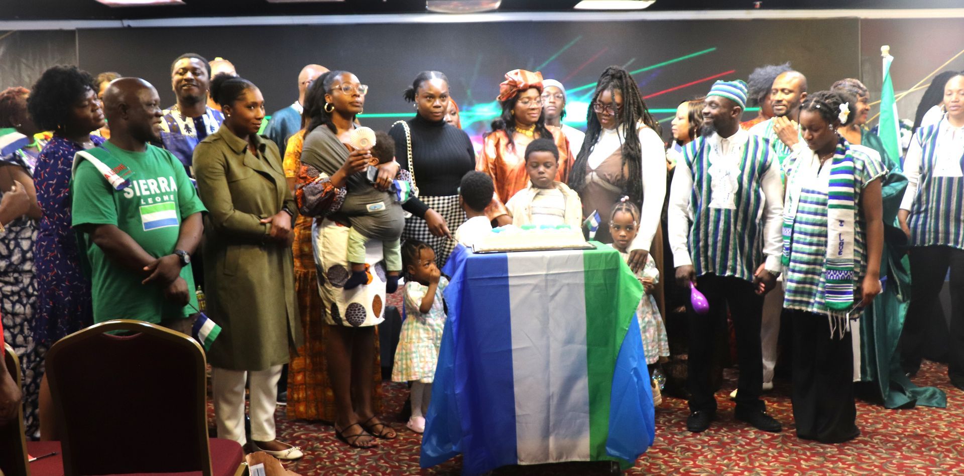 Group of people, some with children, gathered around a table draped in a Sierra Leone flag.