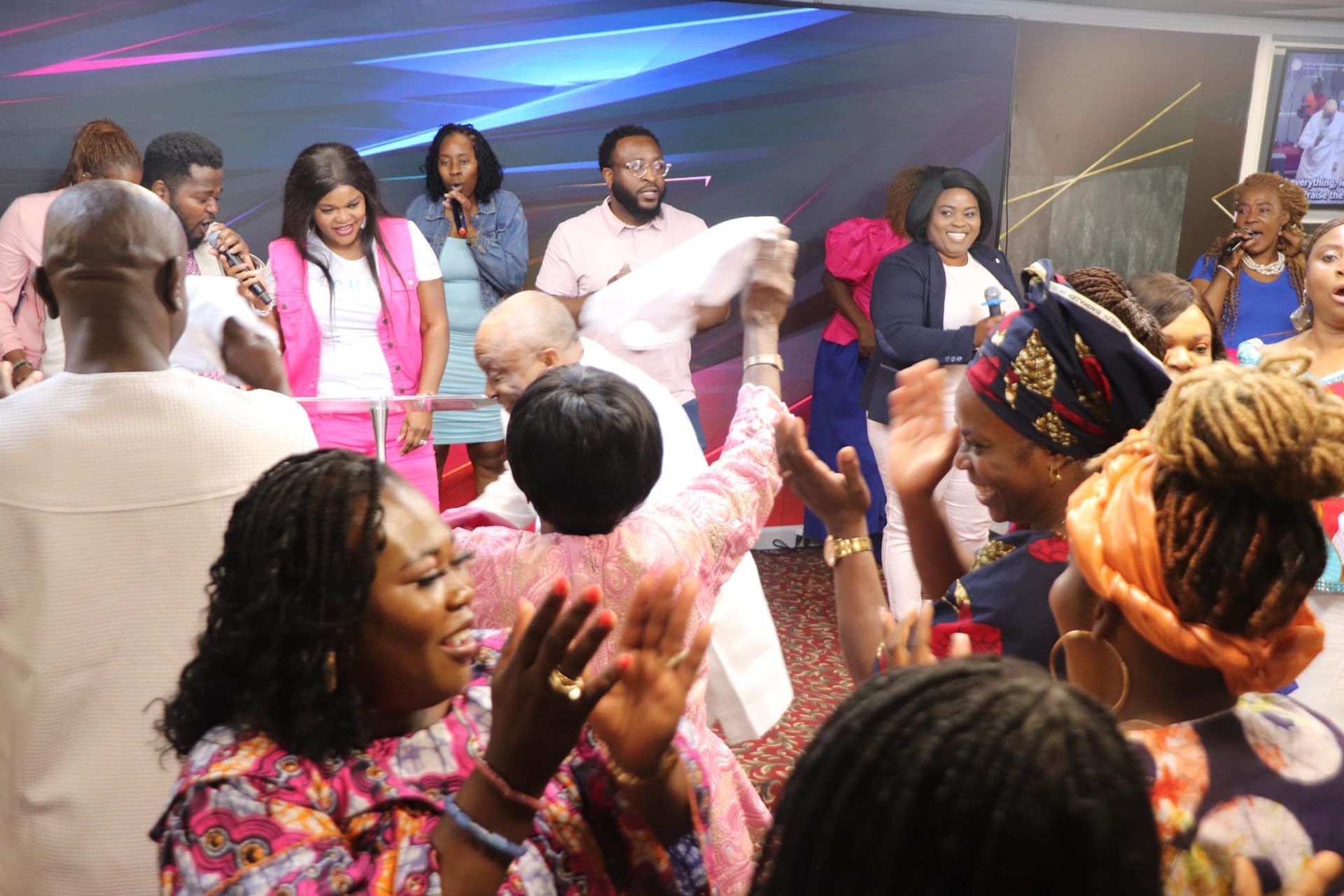 People celebrating in a church. Women in vibrant clothing and accessories. Musicians and a preacher with a white garment.