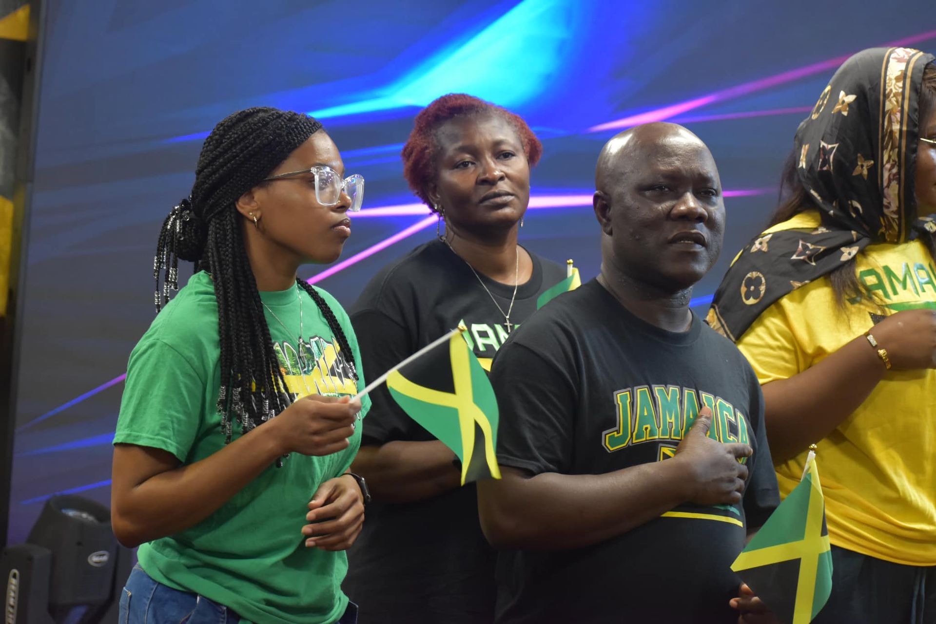 People with Jamaican flags and shirts, some with hands over hearts, indoors.