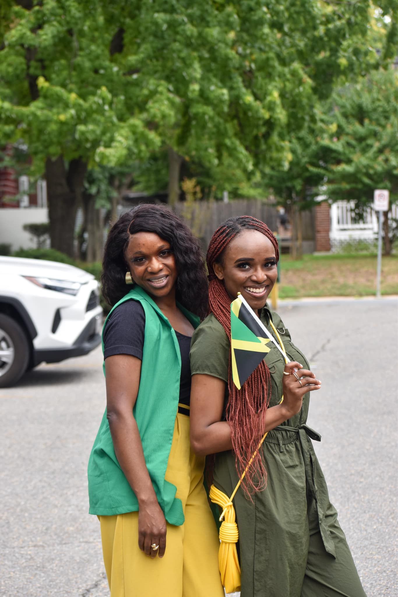 Two women smiling, one holding a Jamaican flag. Trees and a car are in the background.