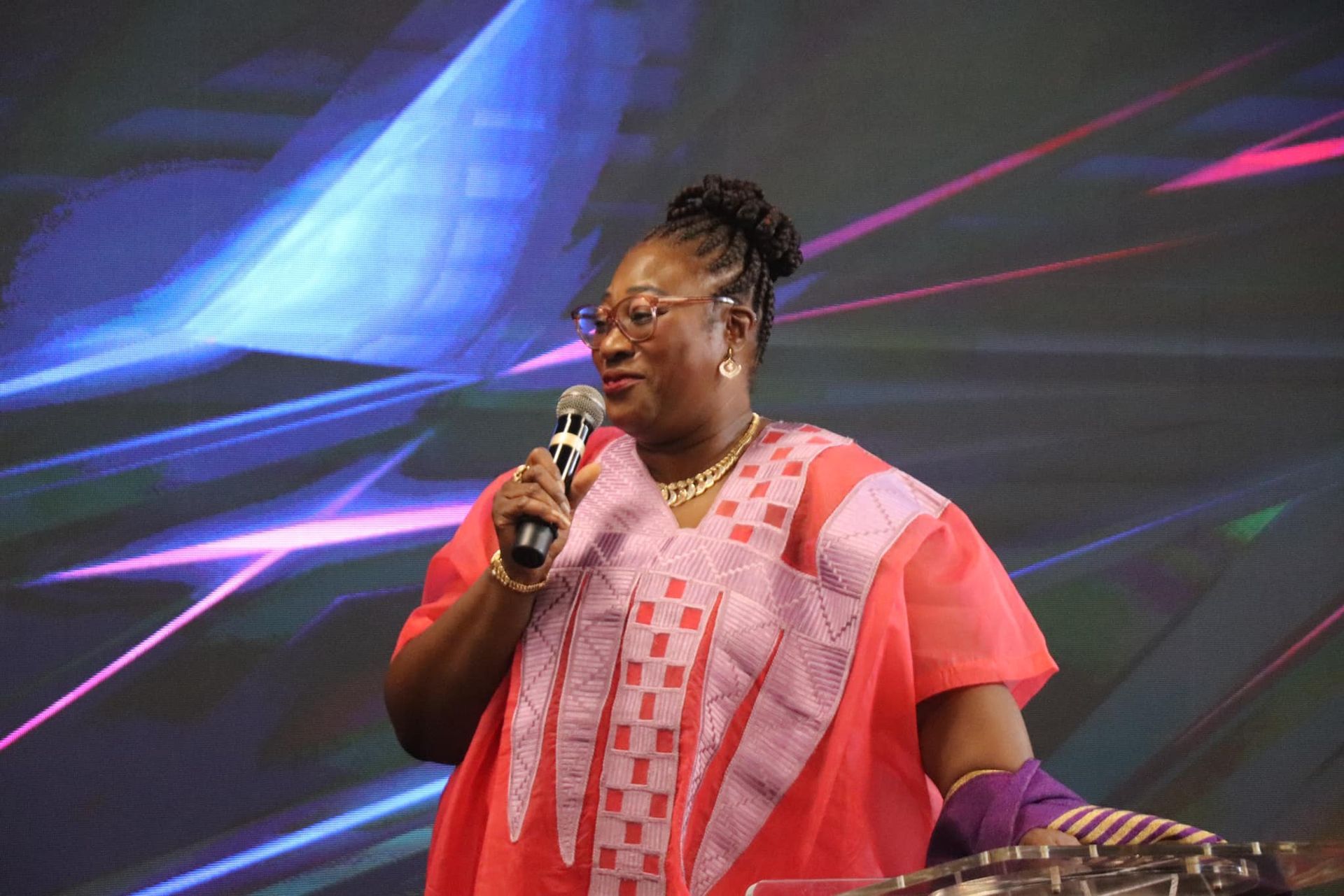 Woman with microphone, in red and purple outfit, speaking on stage with blue and pink light backdrop.