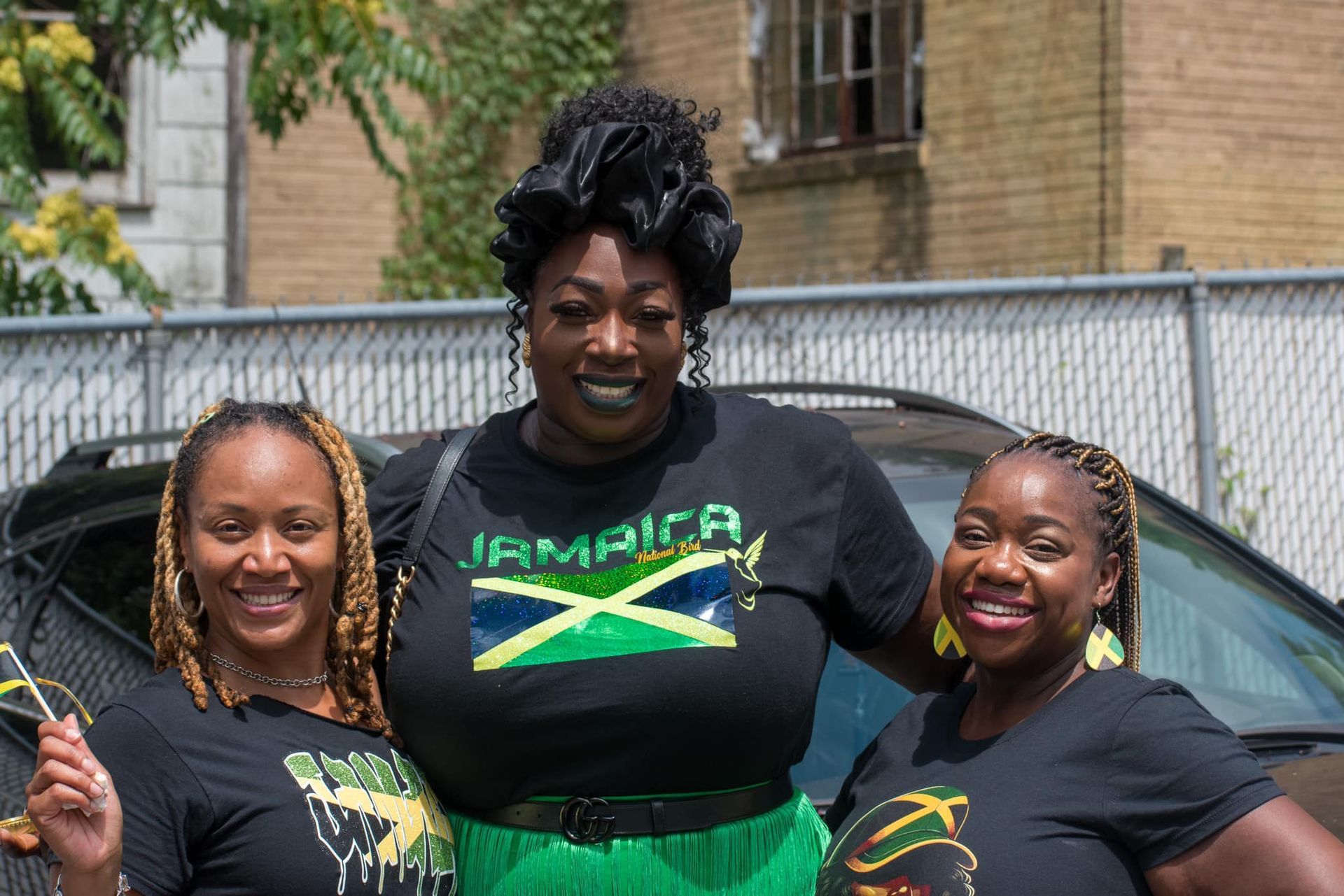 Three women smiling, wearing black shirts with Jamaican flag designs, celebrating outside.