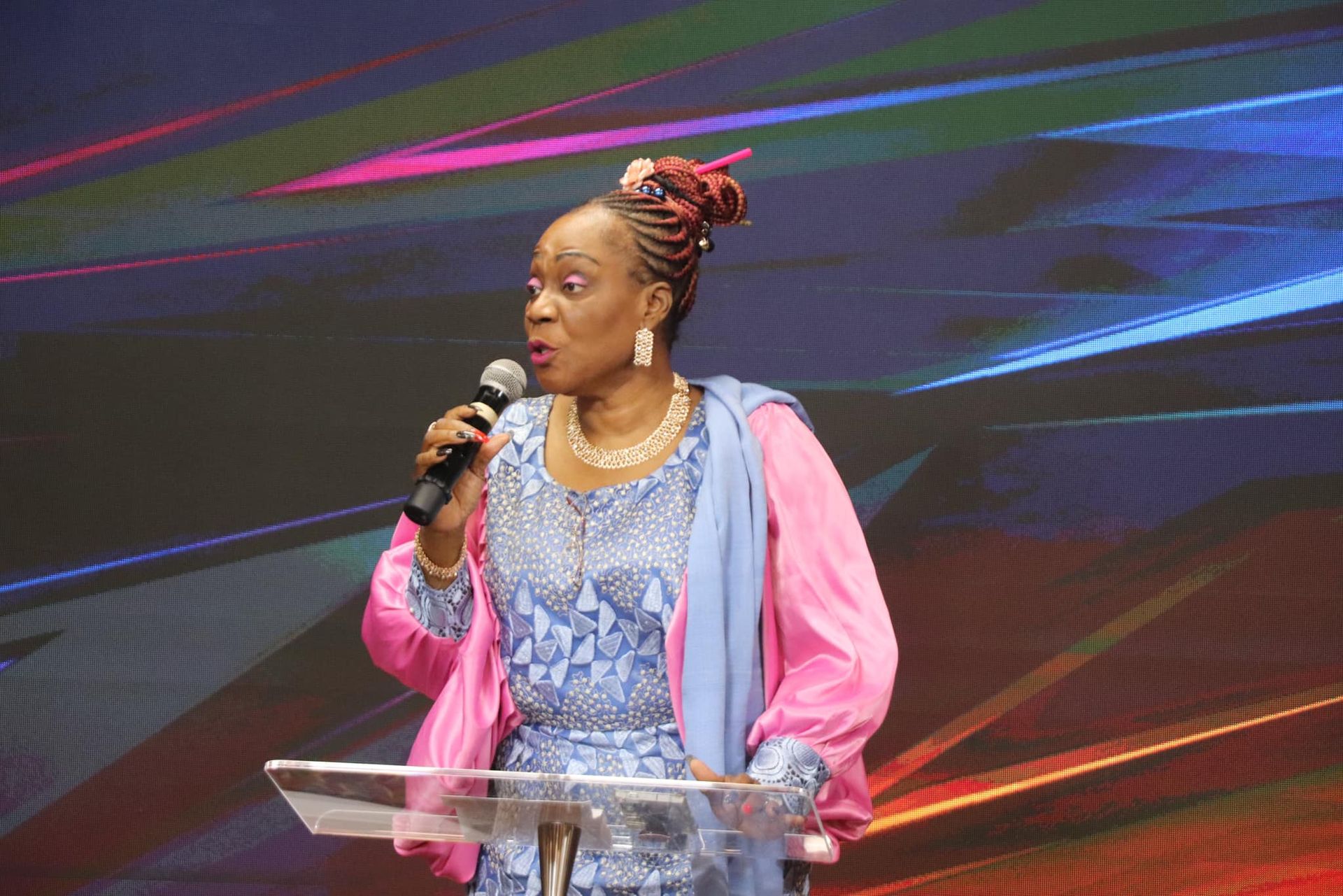 Woman with microphone speaking at a podium, wearing blue dress and pink jacket, stage backdrop.