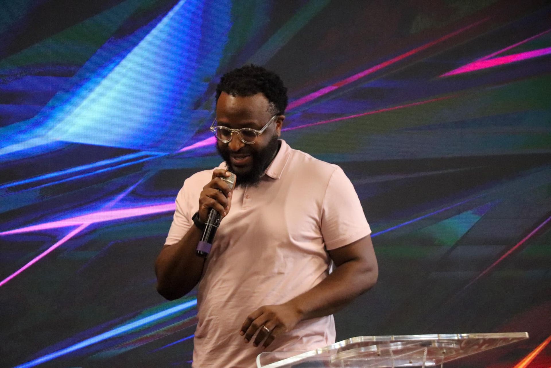 Man speaking into a microphone, standing at a podium, colorful background.