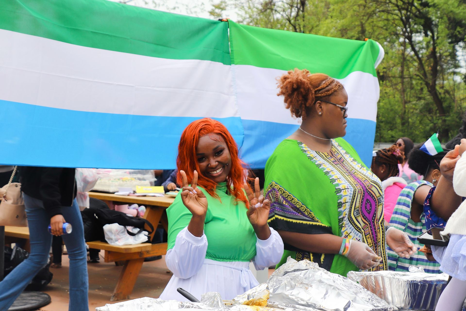 Woman with orange hair gives peace sign in front of Sierra Leone flag at outdoor event.