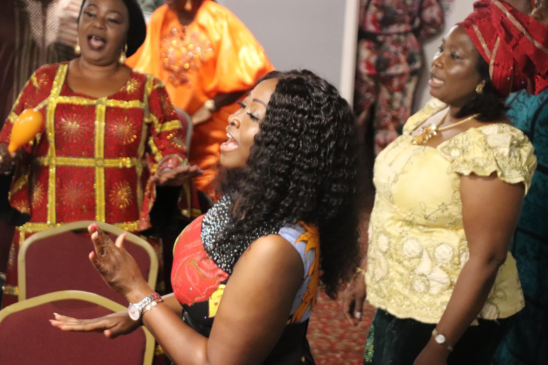 Group of women singing, wearing colorful African attire, in a room.