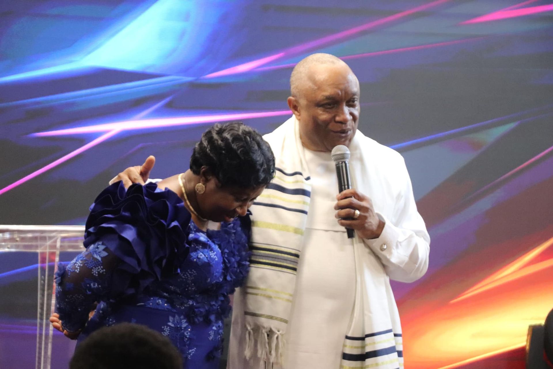 Man in white with a microphone and woman in blue, both bowing at a podium, lit stage background.