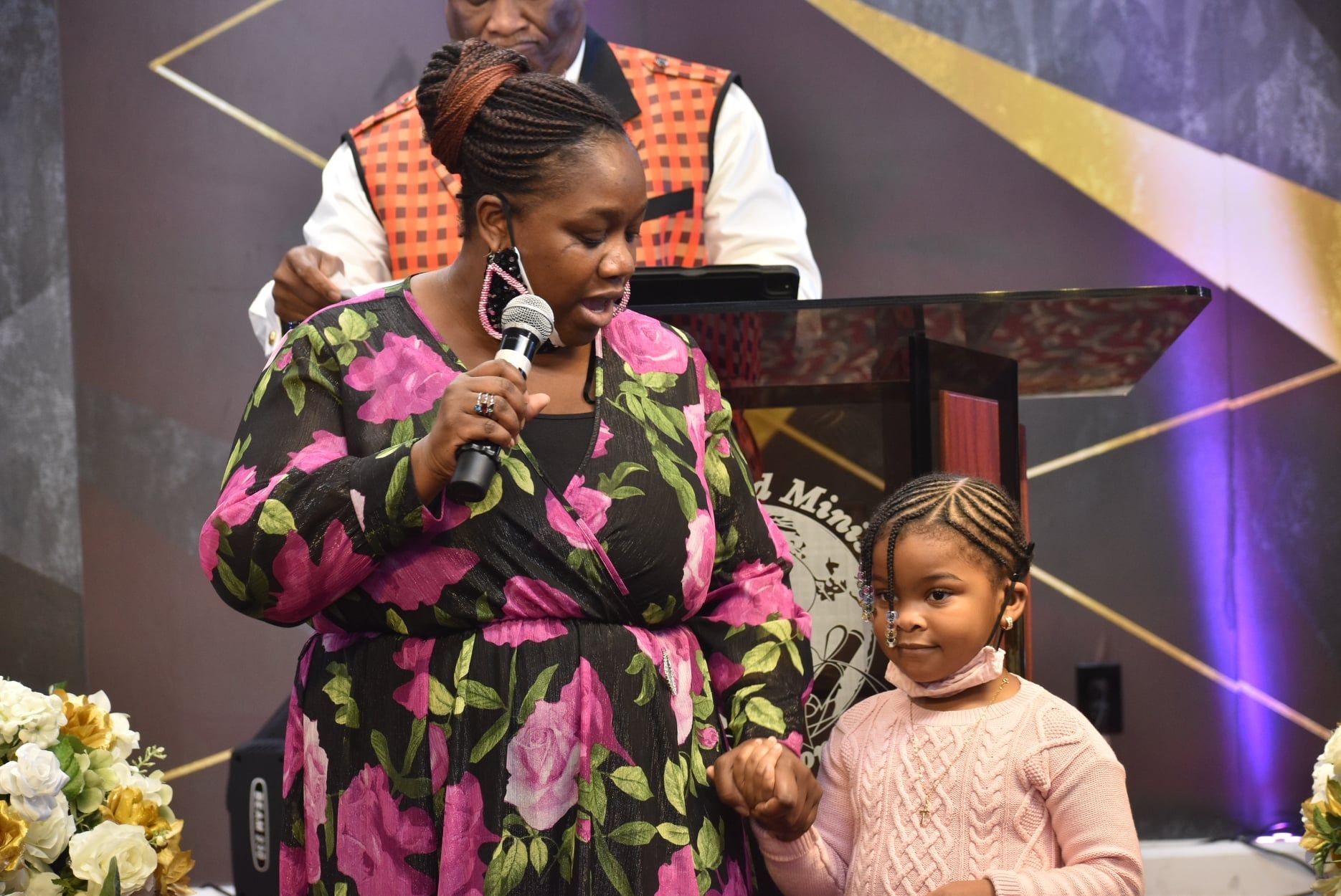 Woman in floral dress speaks into a microphone, holding a young girl's hand onstage at a church.