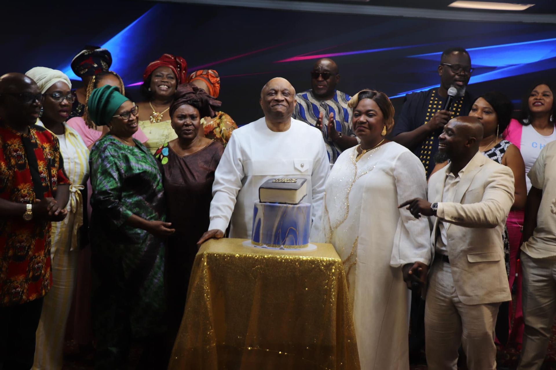 Group of people celebrate around a cake, dressed in various traditional attire. The setting is an indoor event.