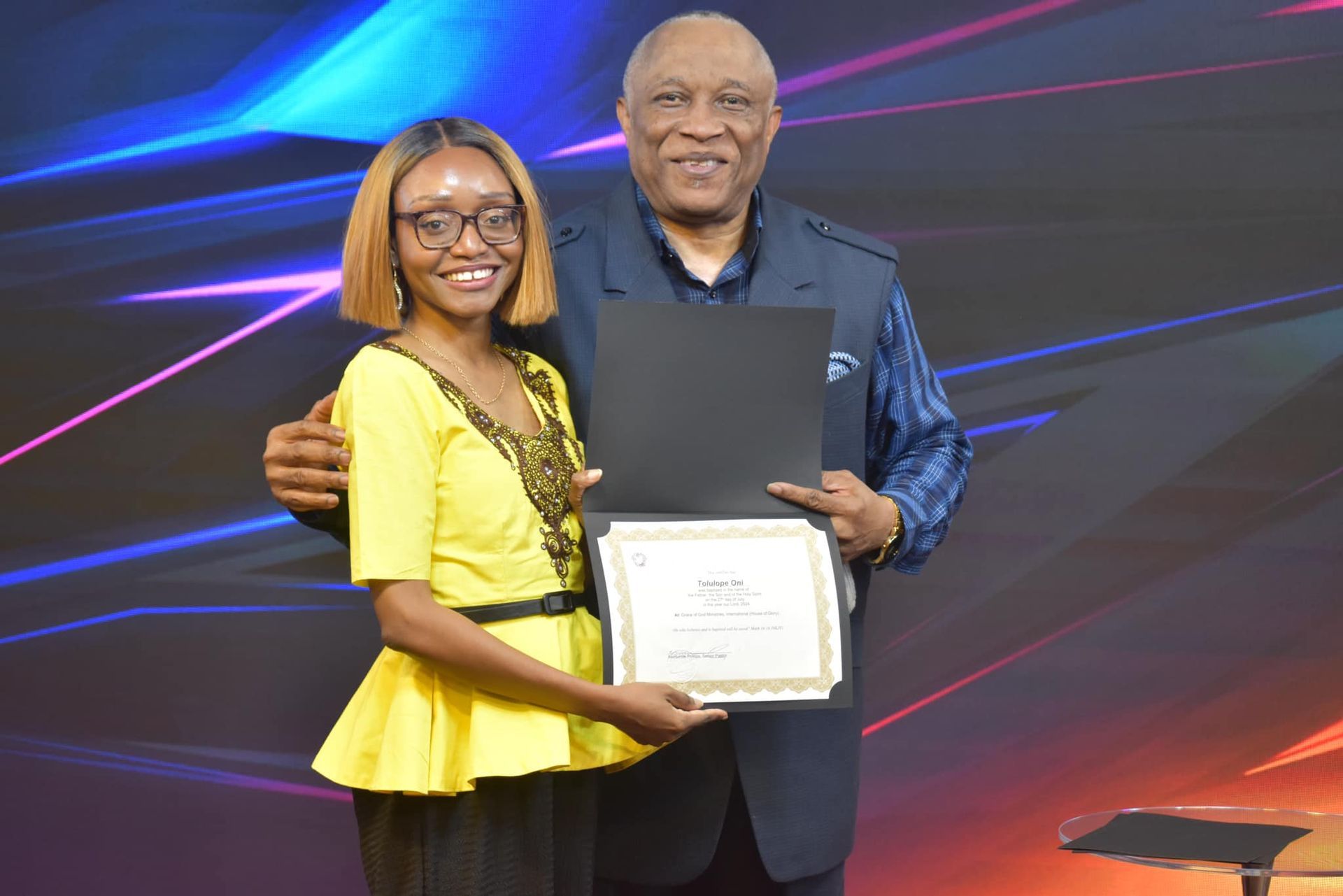 A woman in yellow and a man in blue hold a certificate, smiling, on a stage with colorful lights.