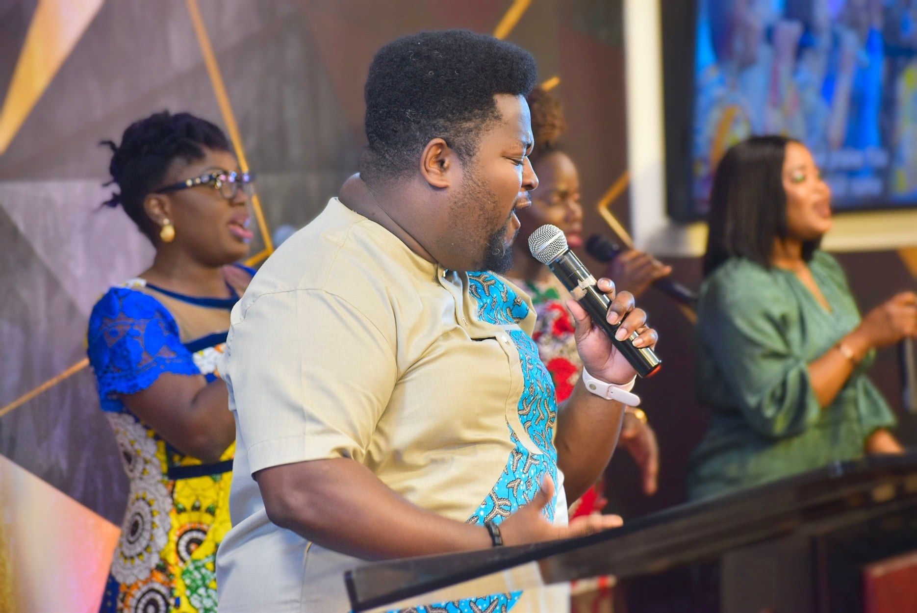A man sings into a microphone on stage, backed by a choir. They appear to be in a church setting.