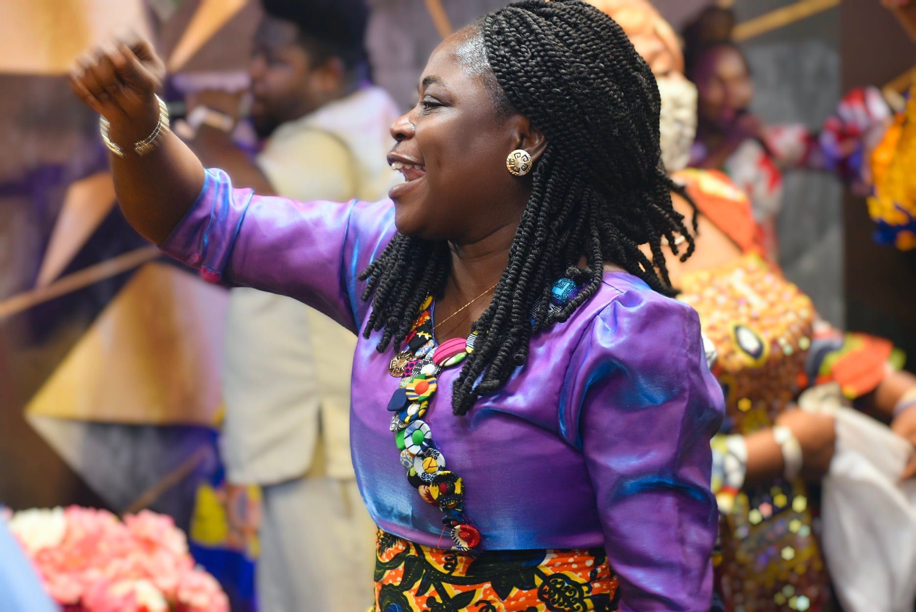 Woman in purple and patterned clothing, smiling and raising arm in a brightly lit setting.