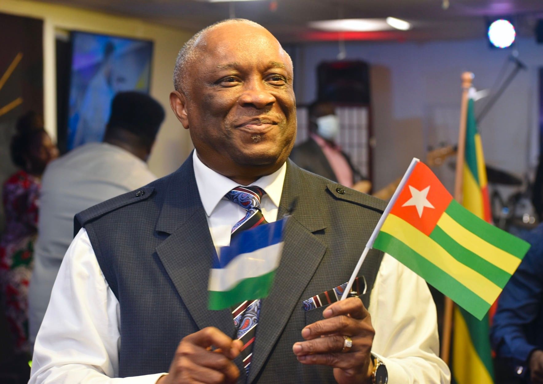 Man in suit holds flags of Sierra Leone and Togo, smiling indoors.