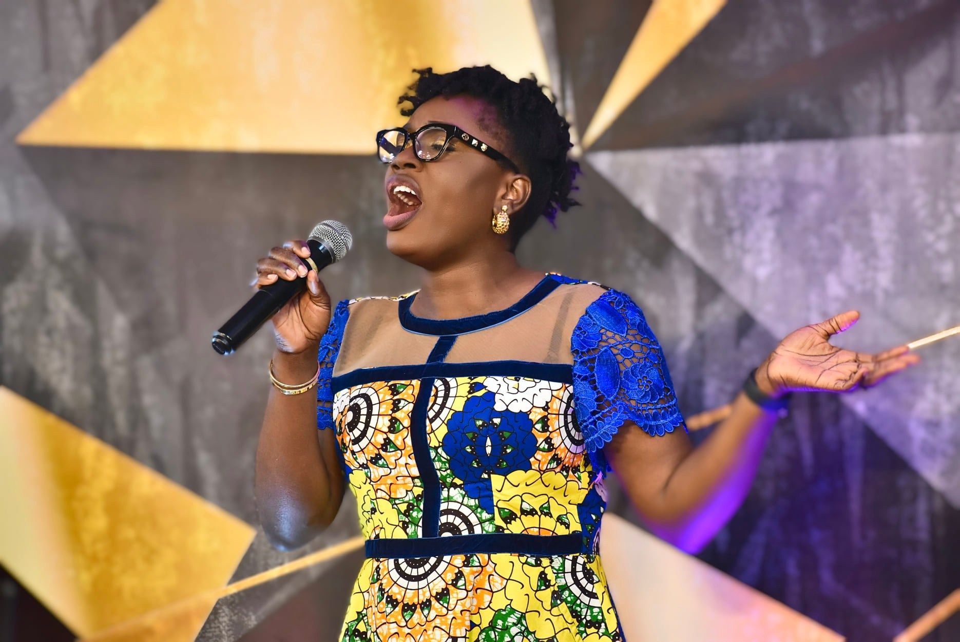 Woman singing on stage, wearing glasses, colorful dress, holding microphone, hands raised.