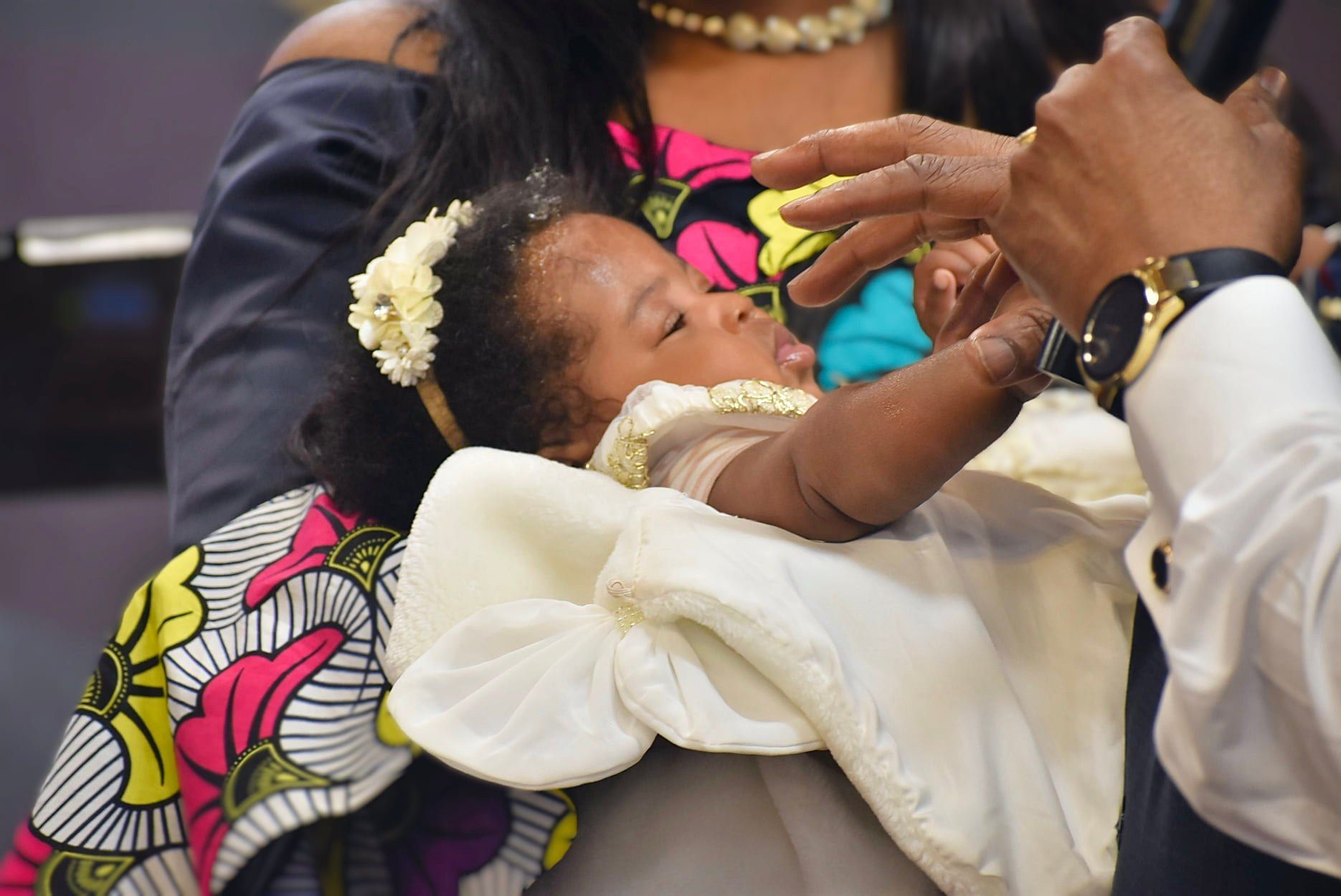 Baby in white gown being fed from a bottle by a person with a gold watch.