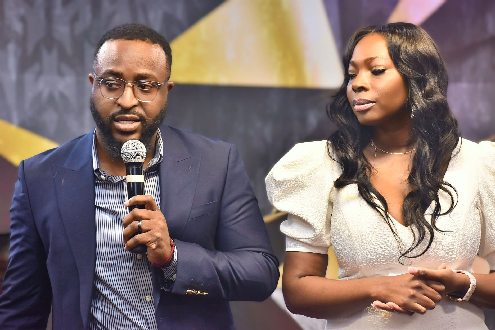 Man speaks into microphone, woman stands beside him. Both are well-dressed. Indoor setting, light colors.