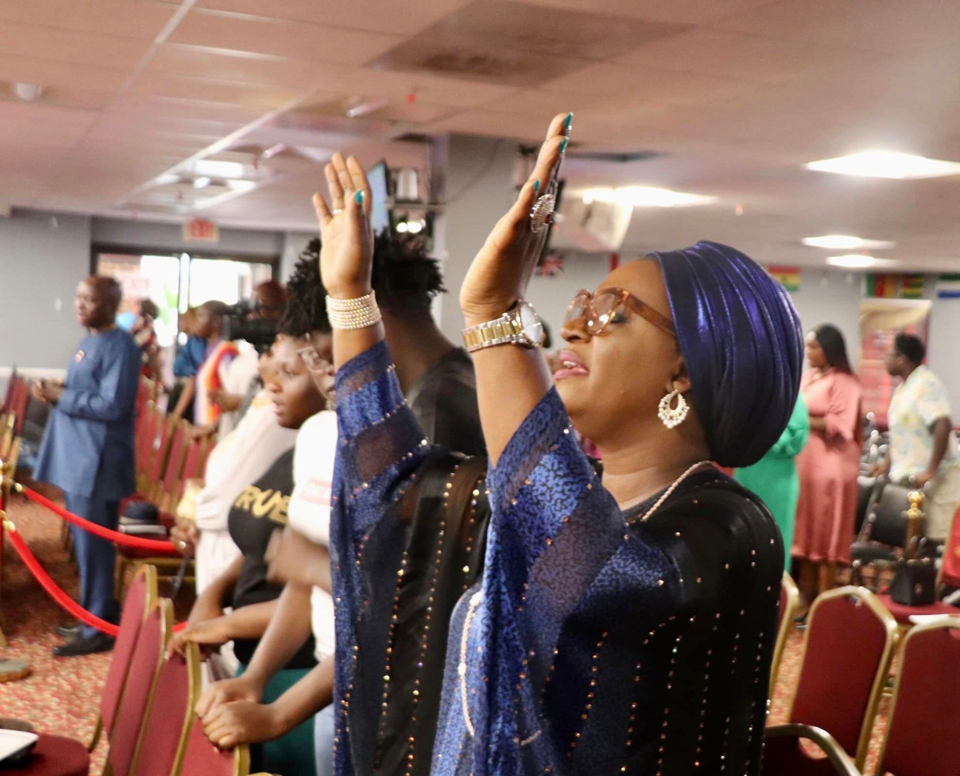 People in a church, some with raised hands. Woman in blue headwrap and dress is praying.